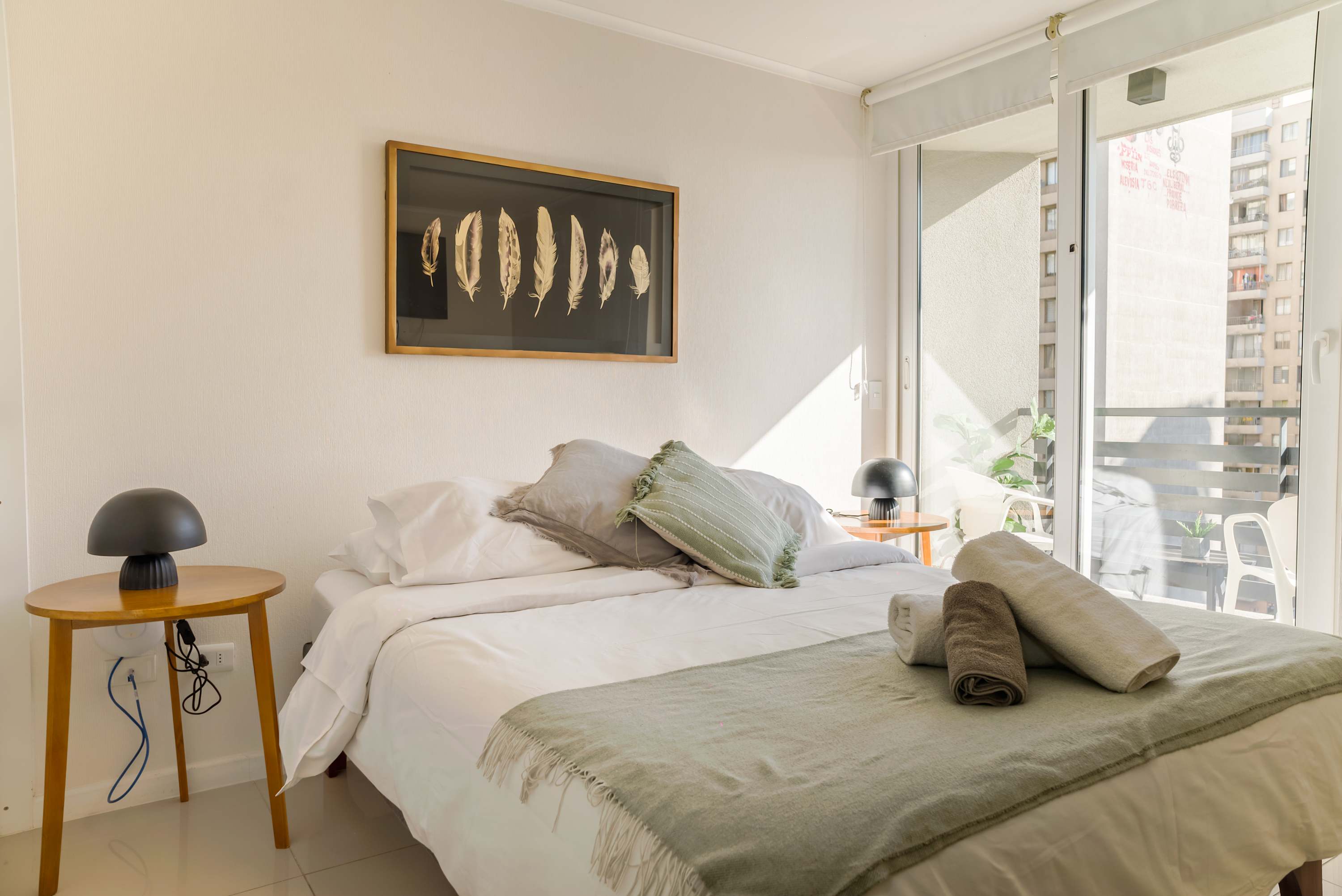 Sunlit studio sleeping area with crisp white bedding, a sage-green throw, feather artwork, and bright balcony doors.