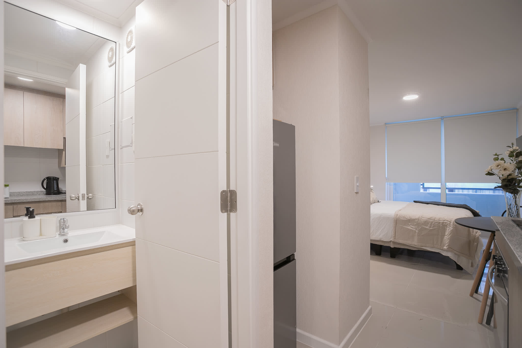 Bright studio interior featuring a neatly made bed, compact kitchenette, and large mirrored bathroom vanity.