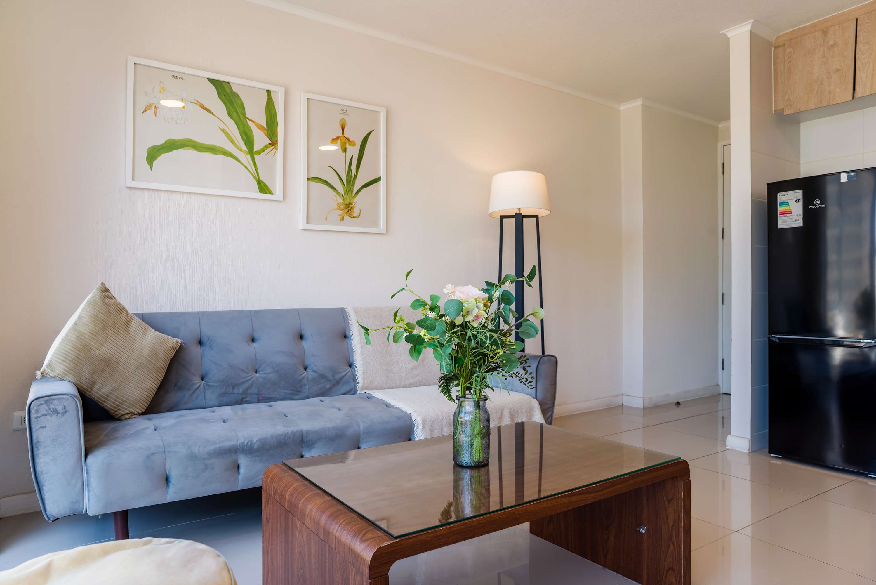 Private living room with a blue-gray sofa, botanical artwork, glass coffee table, and flat-screen TV in a bright studio layout.