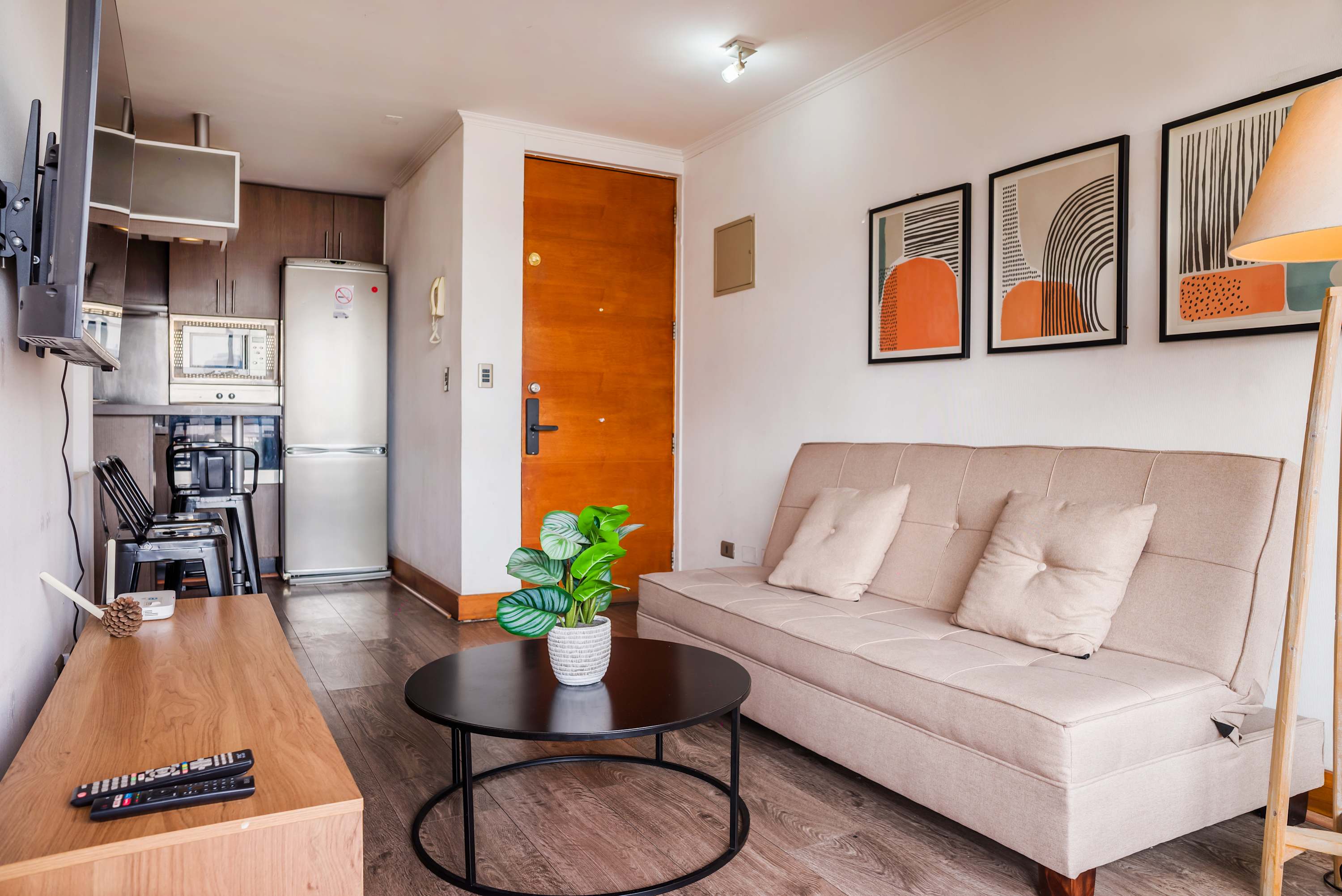 Comfortable living room with a spacious beige sectional, round coffee table, leafy plant, and stylish abstract artwork beside the compact kitchen.