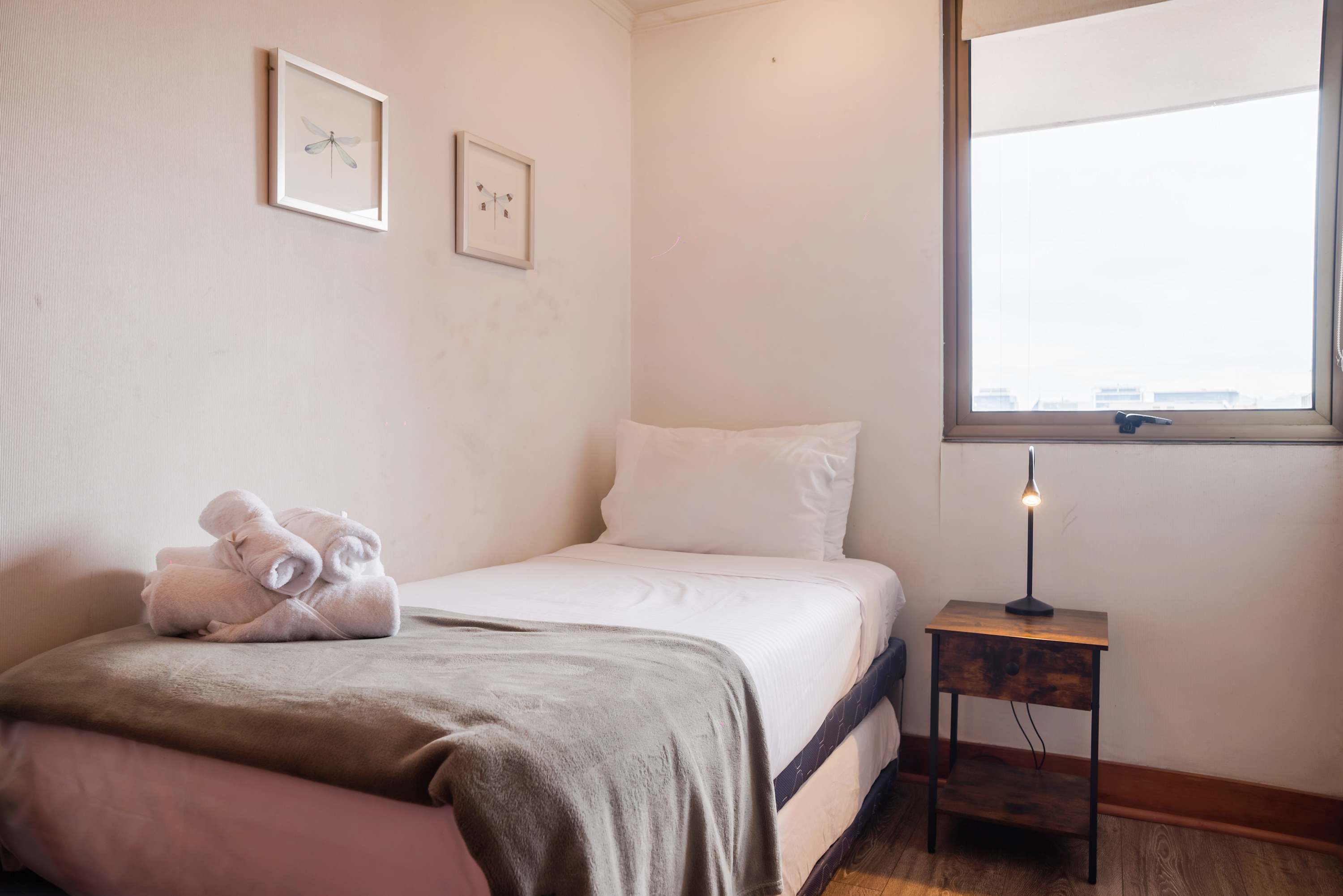 Private bedroom with crisp white bedding, rolled towels, a reading lamp, and a bright city-facing window.