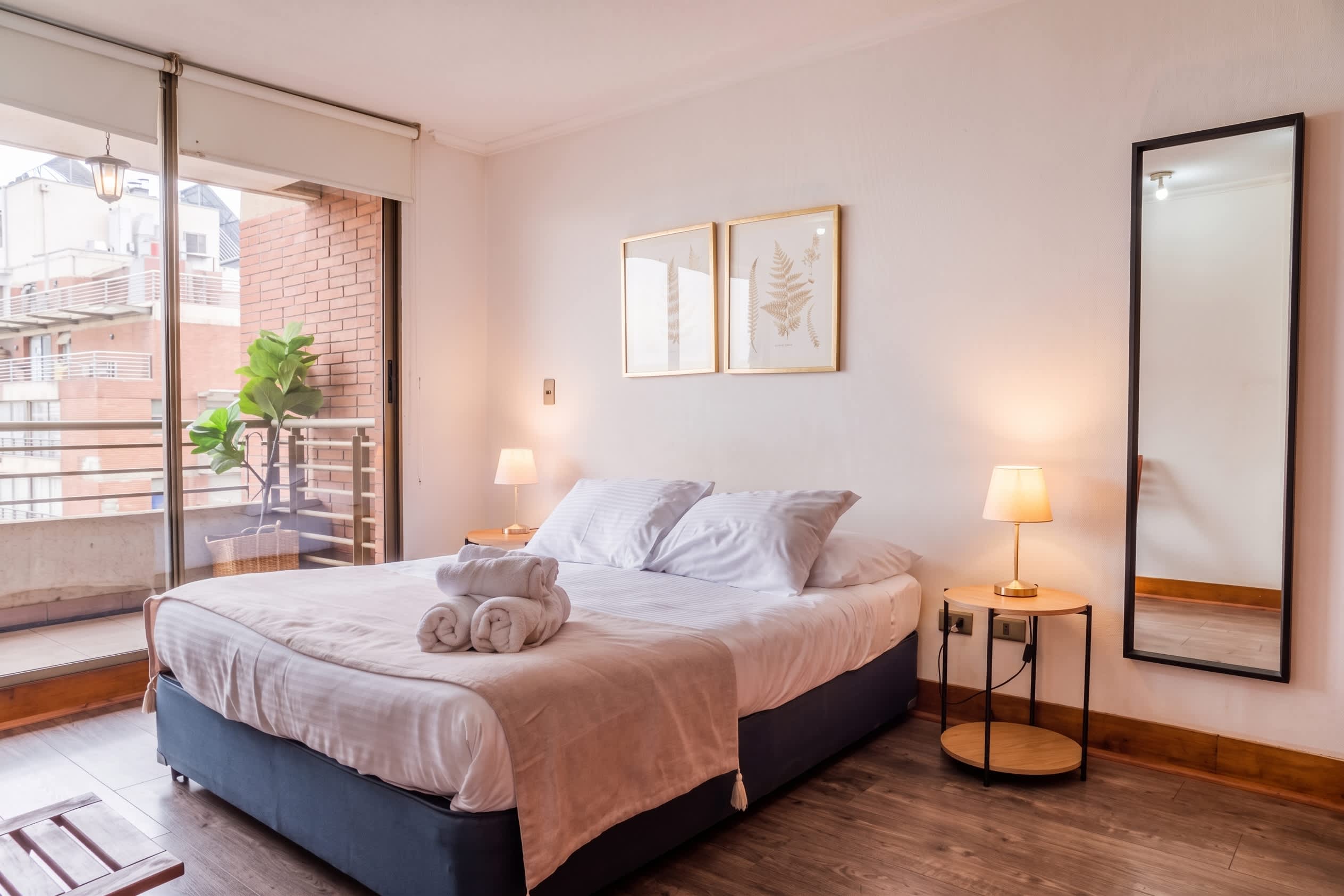 Private bedroom with a neatly made double bed, rolled towels, soft bedside lighting, and glass doors opening to a balcony with a potted plant.