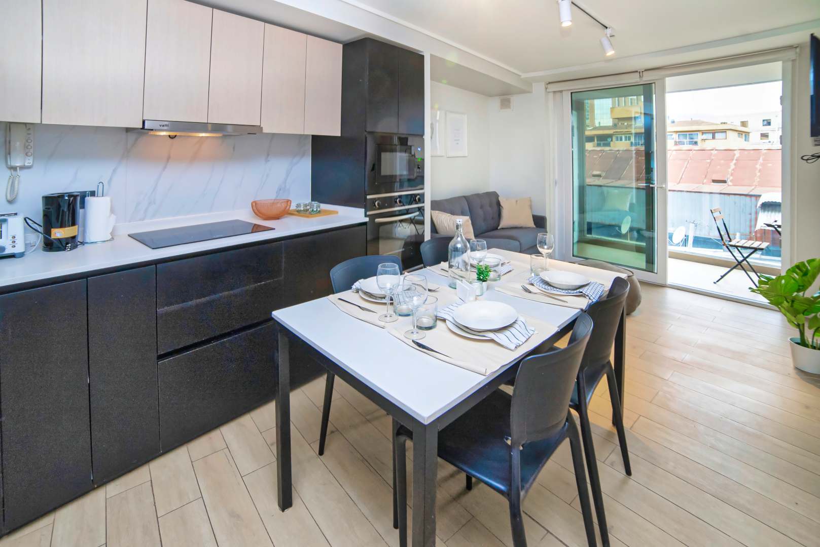 Modern studio kitchen with built-in appliances, a set dining table, and sliding glass doors opening to a balcony with outdoor seating.