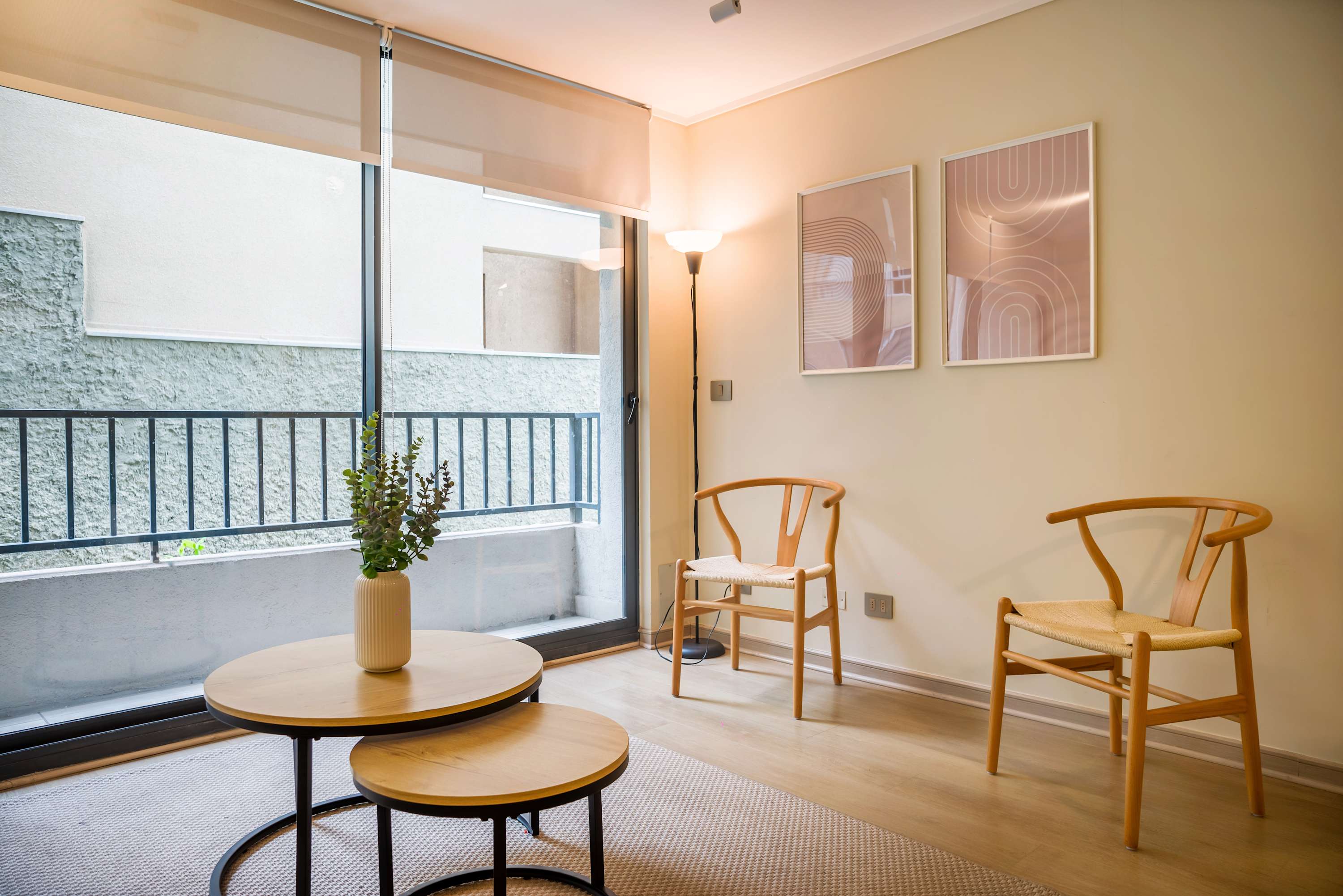 Bright living room with two woven wooden chairs, nesting coffee tables, and floor-to-ceiling windows overlooking the enclosed balcony.