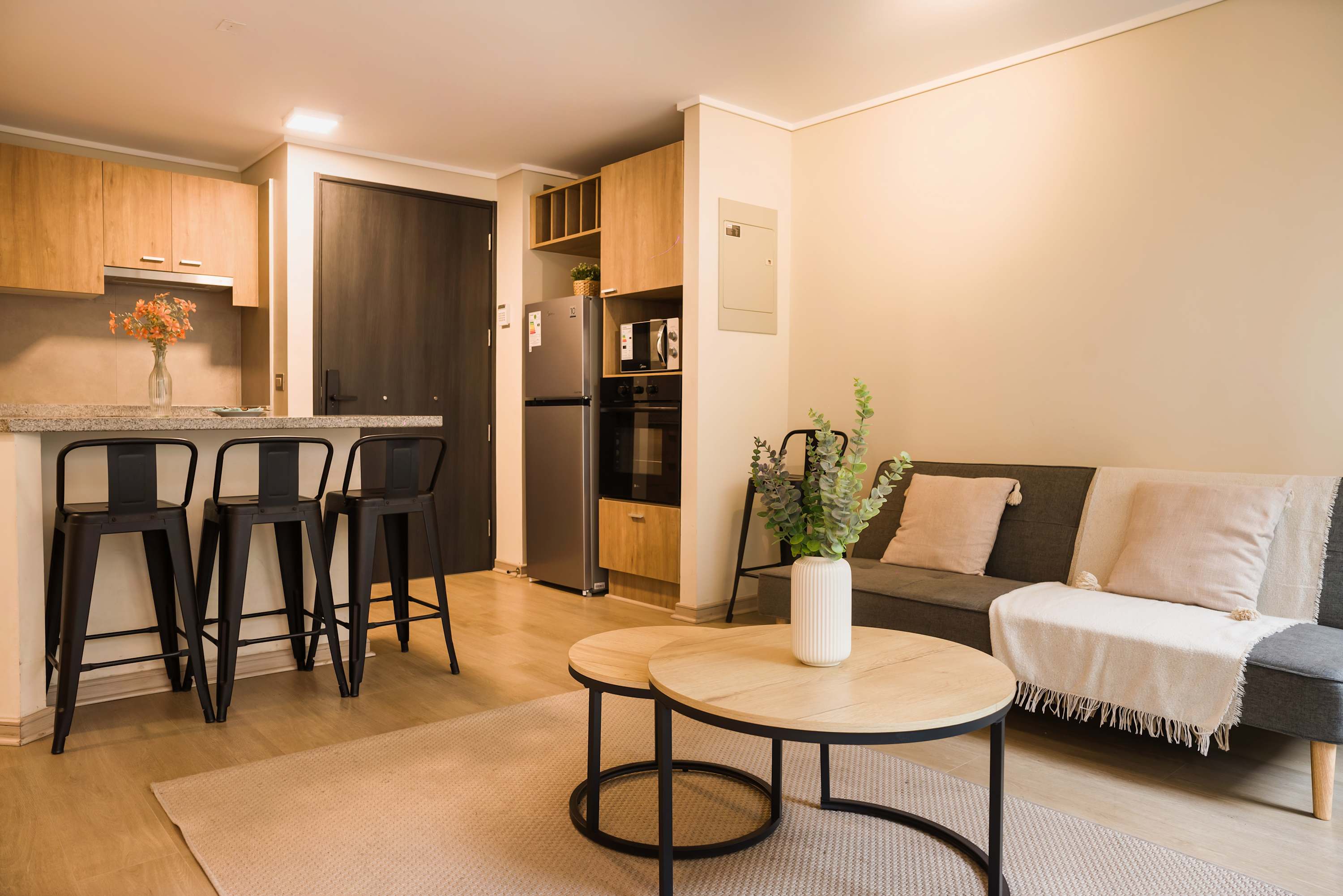 Open-plan living room with a cozy gray sofa, round wood coffee tables, and a fully equipped kitchen with bar seating.