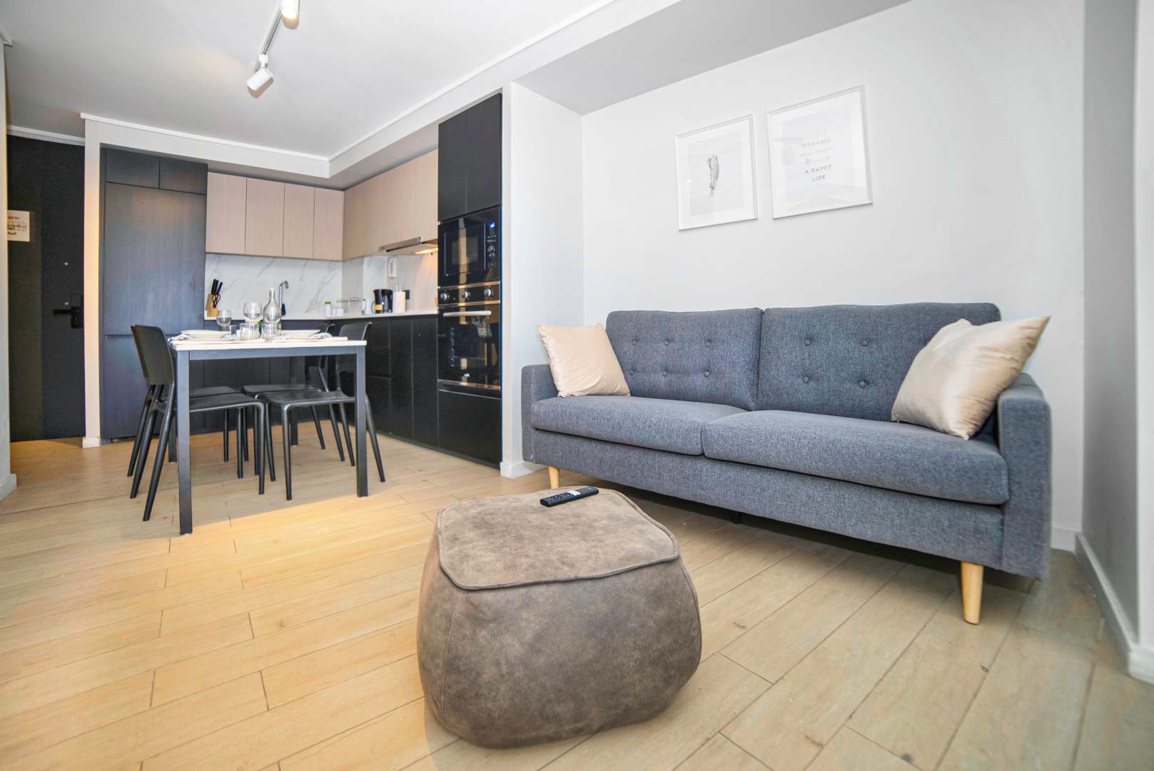 Open-plan living area with a comfortable gray sofa, cushioned ottoman, four-seat dining table, and sleek integrated kitchen.