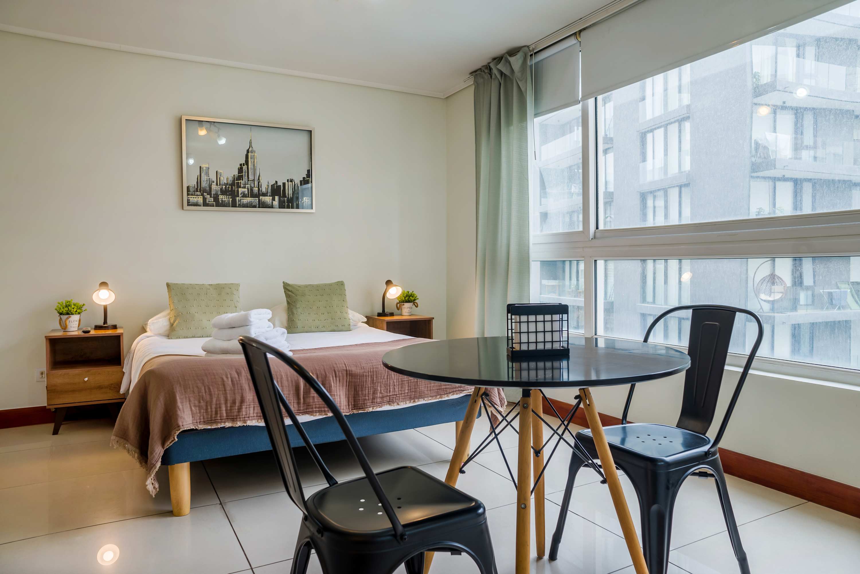 Bright studio living area with a styled double bed, round dining table, and expansive corner windows overlooking neighboring city architecture.
