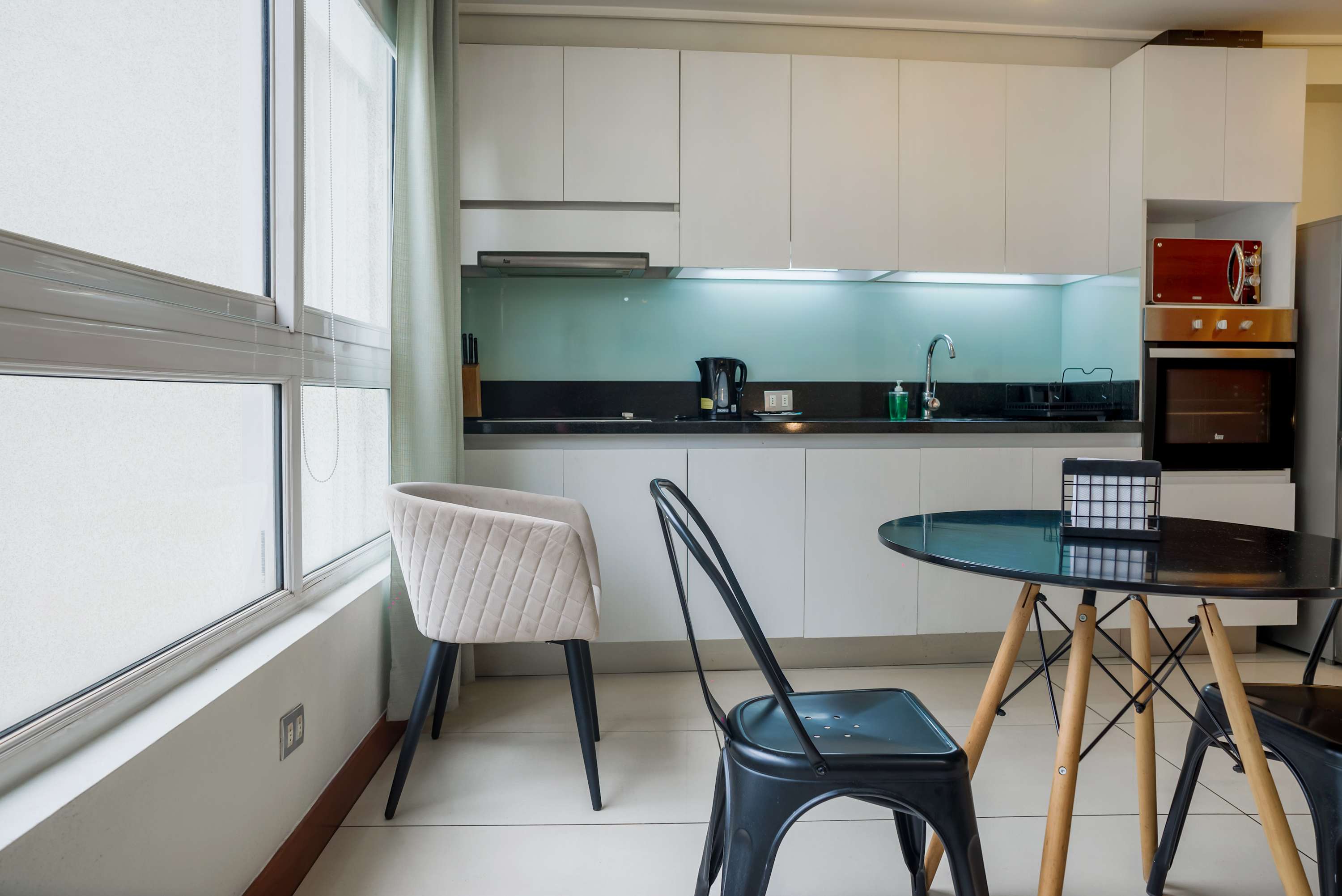 Bright studio kitchen with sleek white cabinetry, a pale turquoise backsplash, and a round dining table beside large windows.