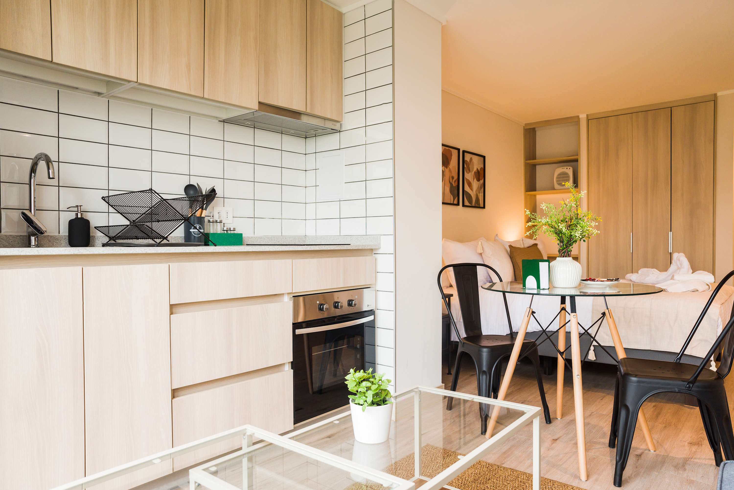 Bright studio kitchen with light wood cabinetry, a tiled backsplash, and a glass dining table beside the comfortable sleeping area.