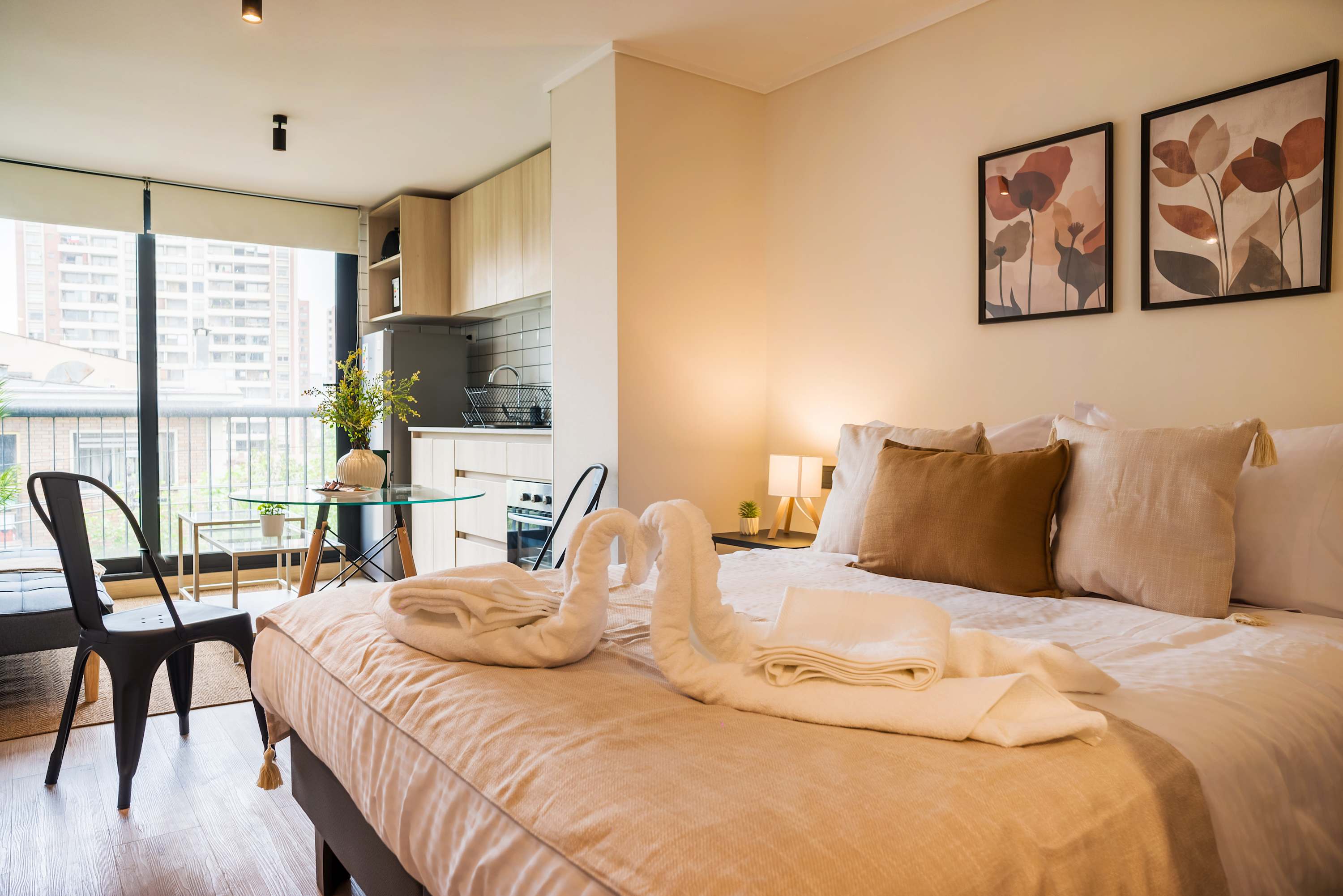 Cozy studio sleeping area with a beautifully dressed bed, compact kitchen, glass dining table, and bright city-facing windows.