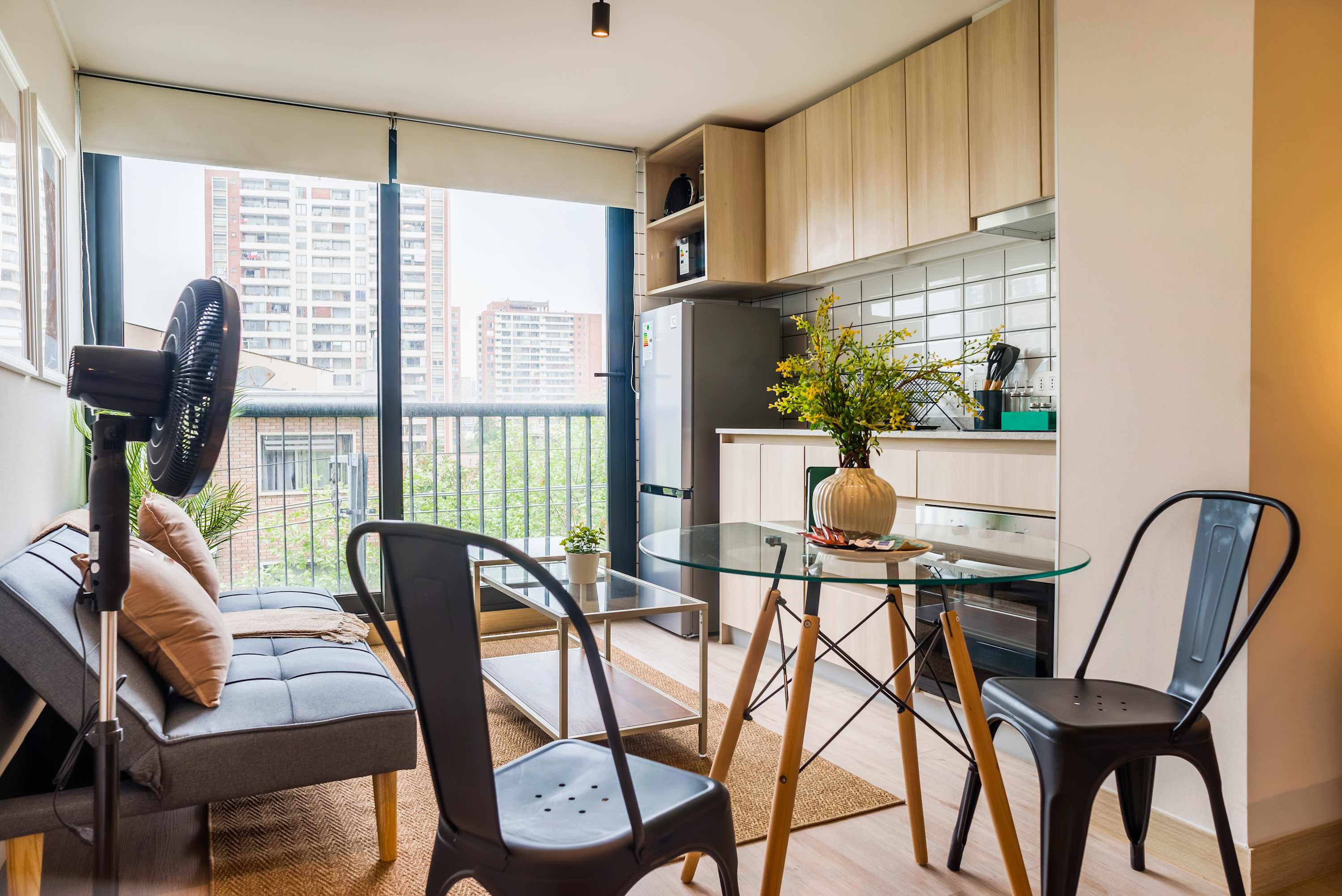 Bright studio living area with a glass dining table, cozy gray sofa, and expansive windows overlooking the city.