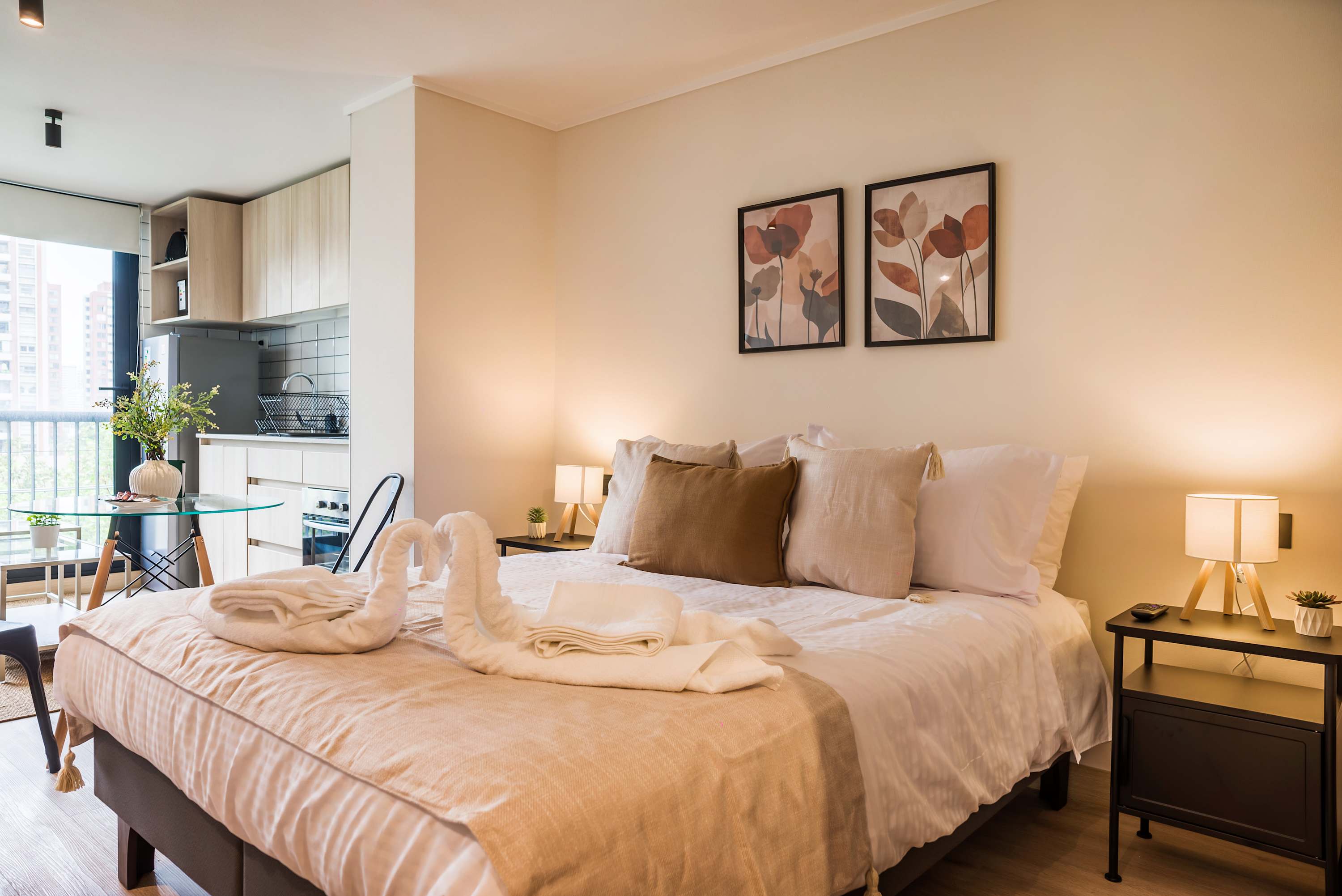 Warm studio sleeping area with layered bedding, botanical artwork, and a compact kitchen beside a bright city-facing window.