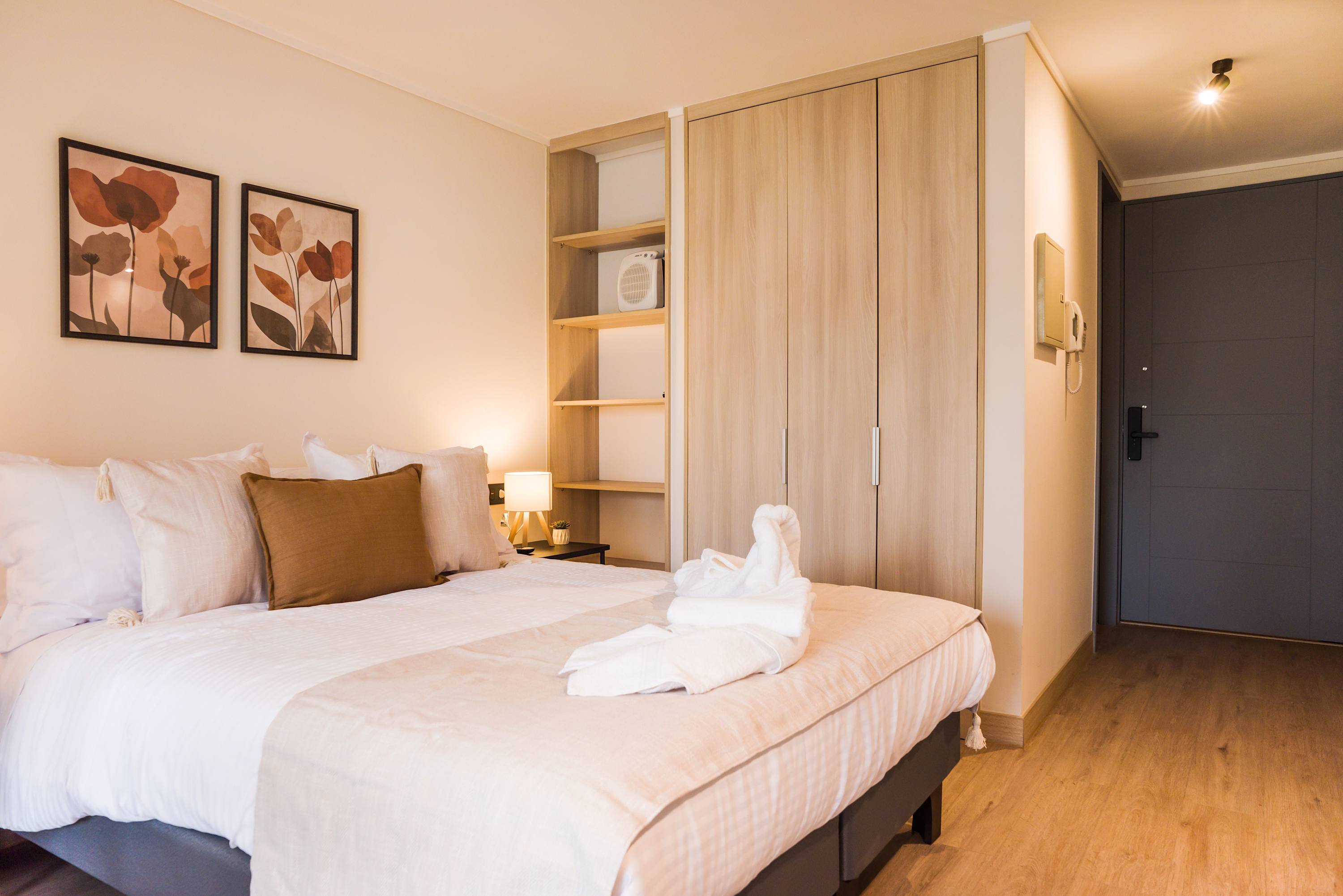Studio sleeping area with crisp white bedding, soft beige accents, botanical artwork, and a light-oak built-in wardrobe.
