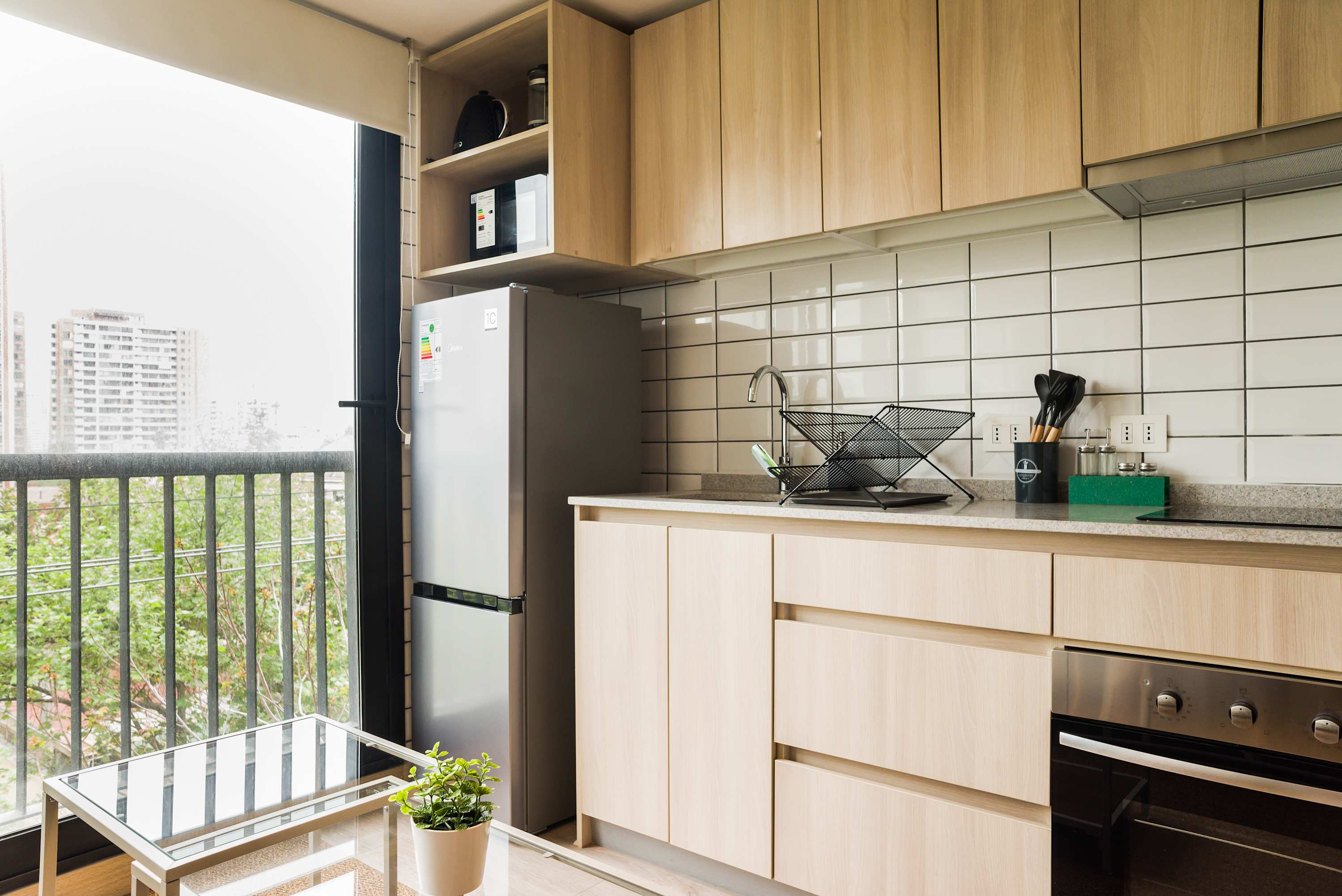 Bright studio kitchen with light wood cabinetry, full-size appliances, and a leafy balcony outlook.
