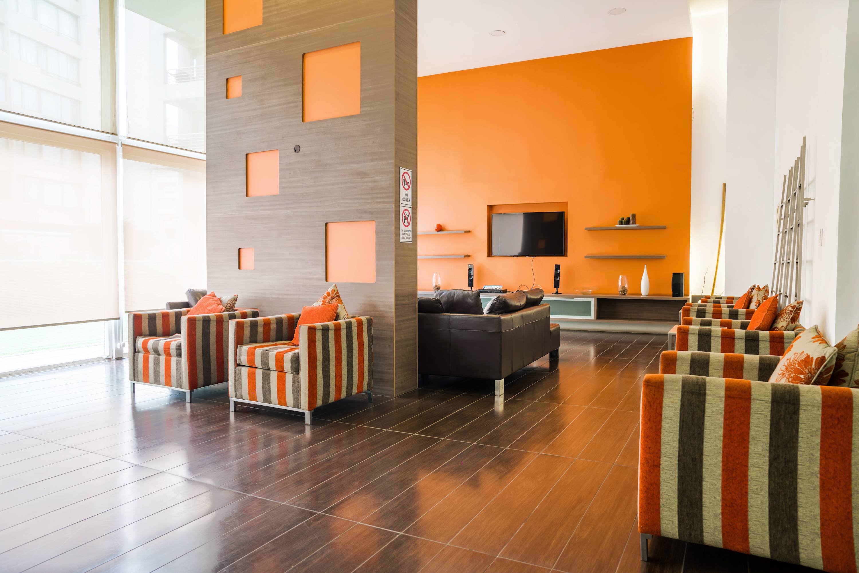 Bright shared lounge with floor-to-ceiling windows, bold orange accents, comfortable striped armchairs, leather sofas, and a wall-mounted TV.