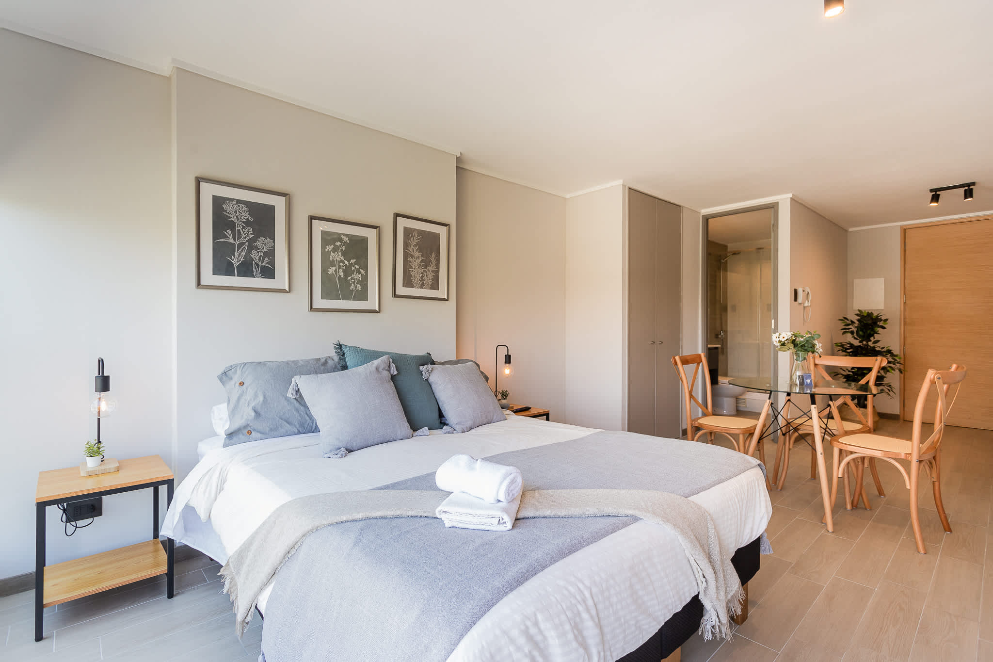 Bright studio retreat with a neatly styled double bed, soft blue-gray textiles, botanical artwork, and a dining area beside the glass-enclosed bathroom.