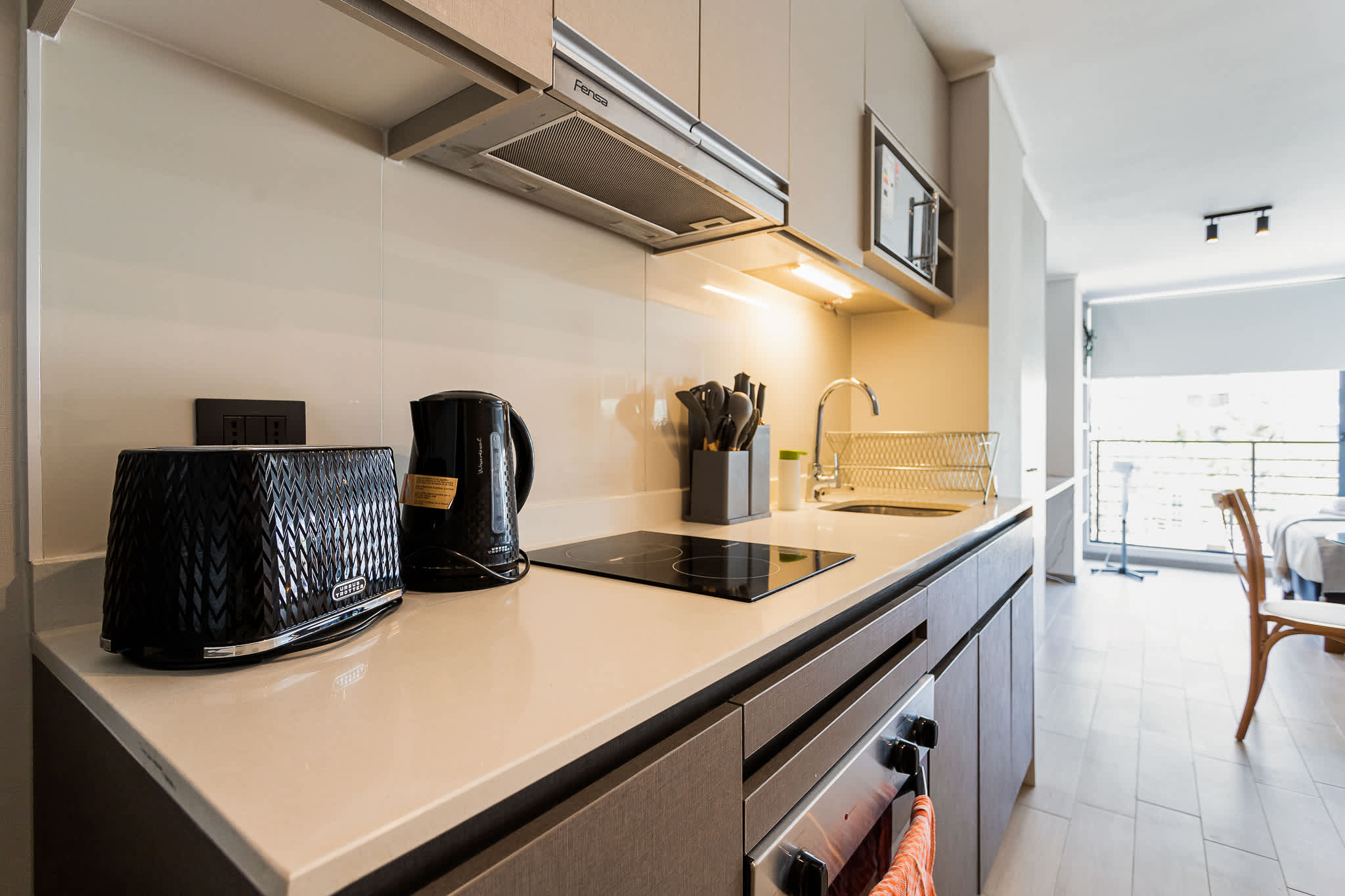 Modern studio kitchen with a sleek induction cooktop, built-in appliances, and a bright open-plan dining area beyond.