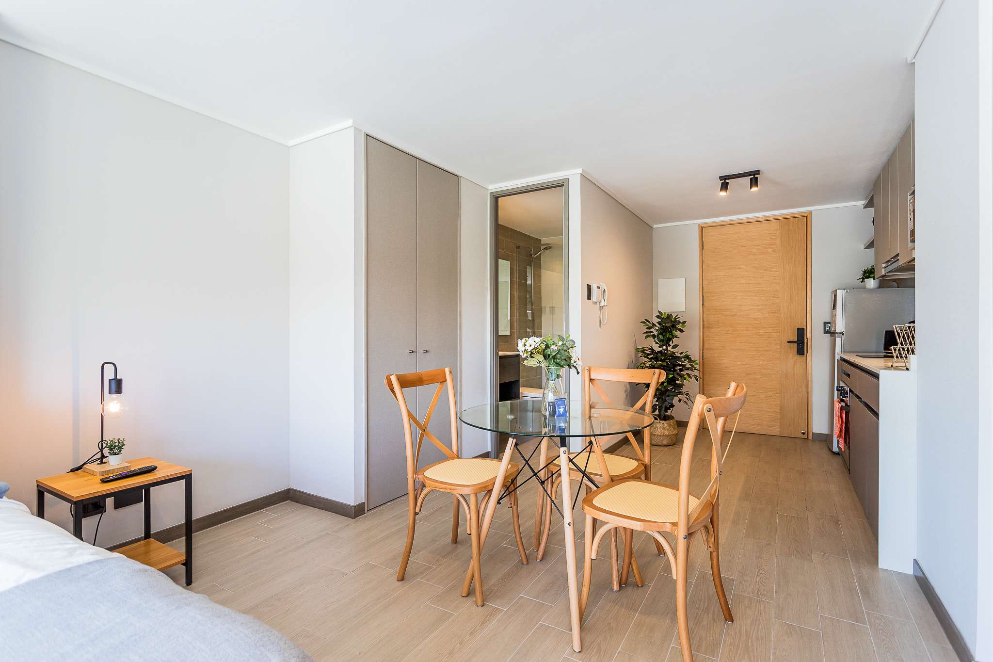 Open-plan studio dining area with a round glass table, four wooden chairs, and a compact kitchen nearby.