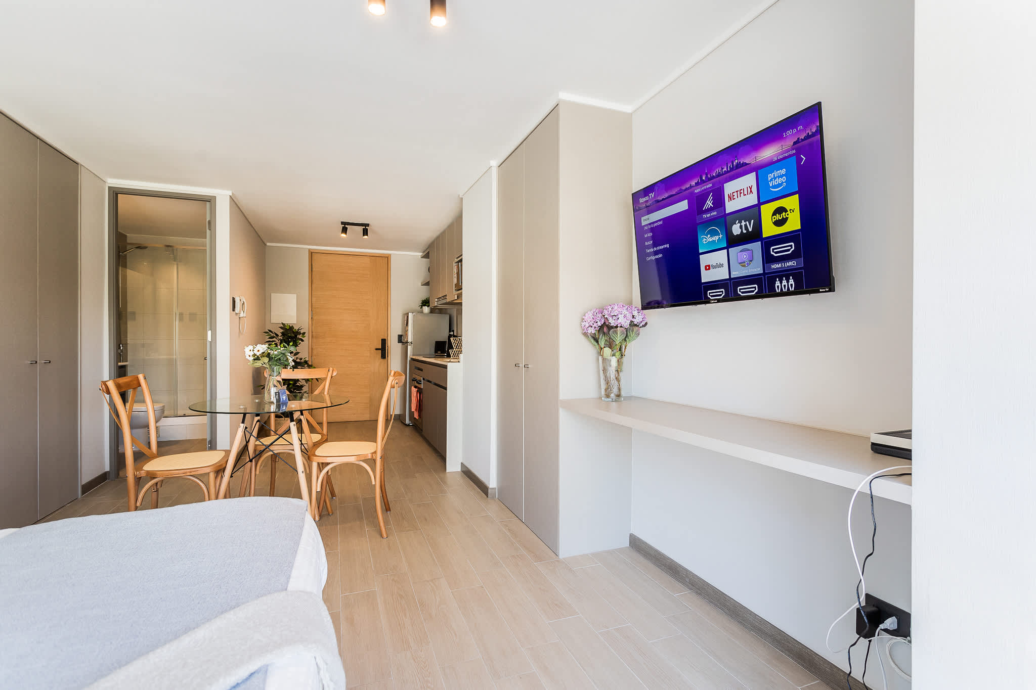 Bright studio living area with a wall-mounted smart TV, round glass dining table, and warm wood finishes.