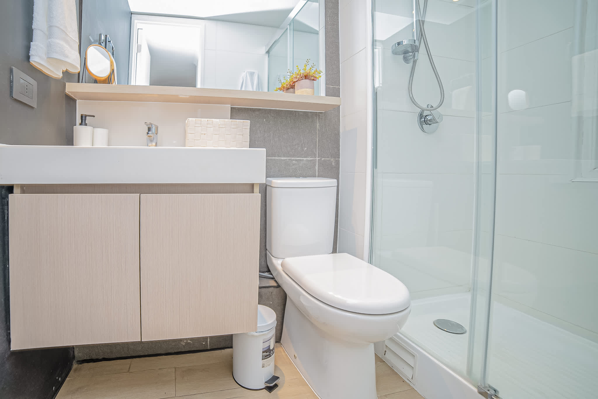 Bright private bathroom with a glass-enclosed shower, modern vanity, chrome fixtures, and soft wood-and-gray finishes.