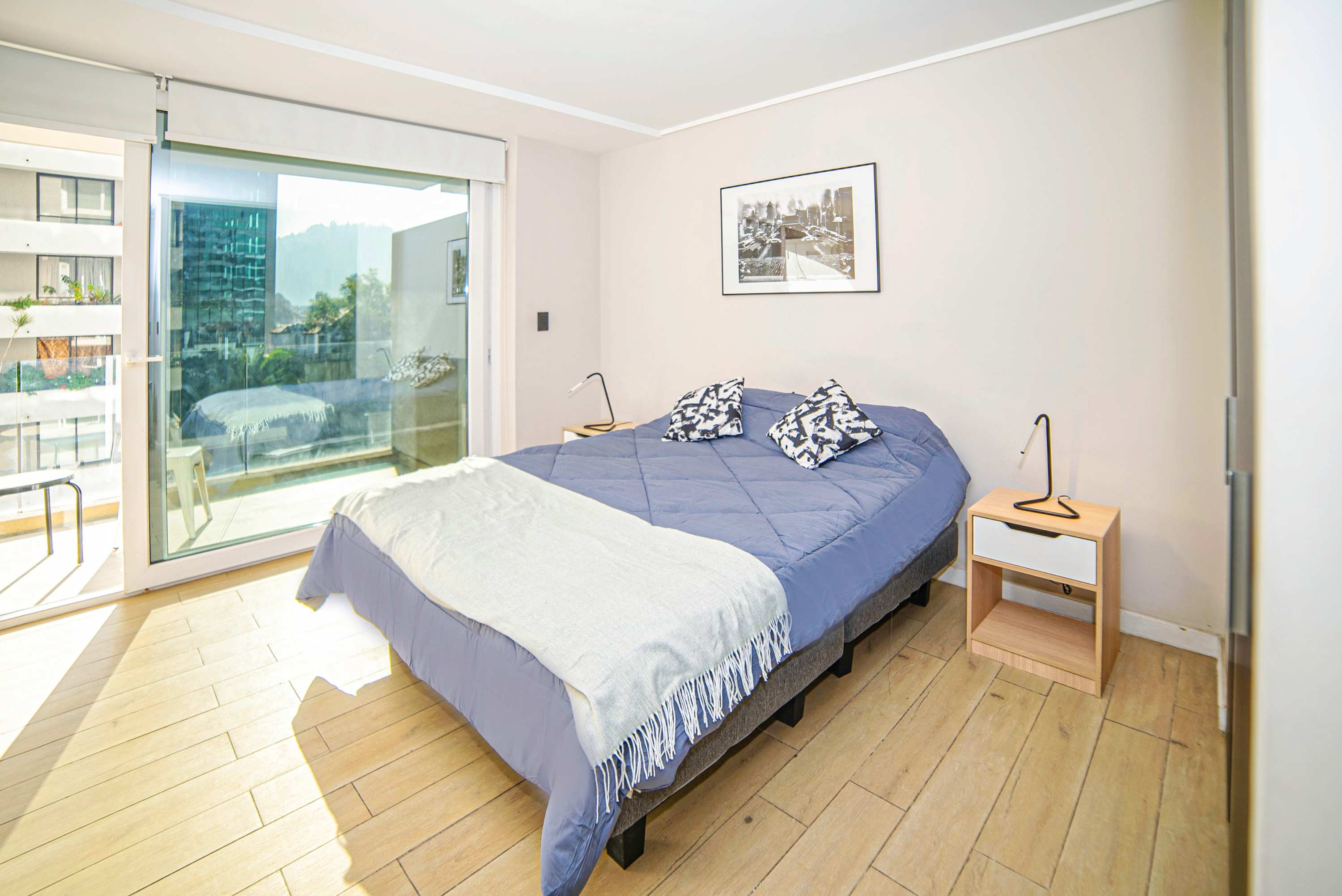 Studio sleeping area with a blue quilted bed, minimalist bedside lamps, and bright glass doors opening to a balcony view.