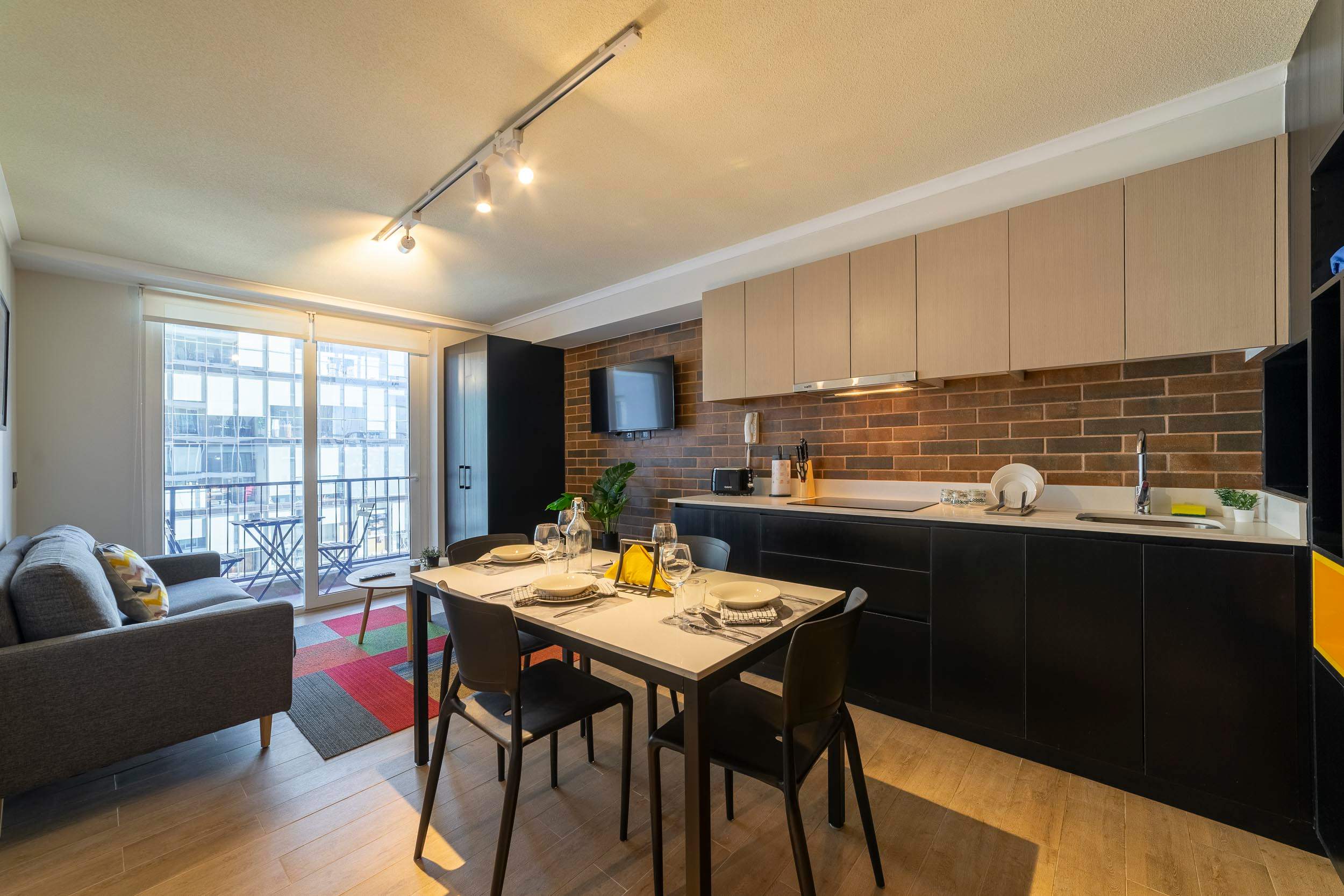 Open-plan living and dining room with a four-seat table, sleek black-and-wood kitchen, and bright balcony doors.