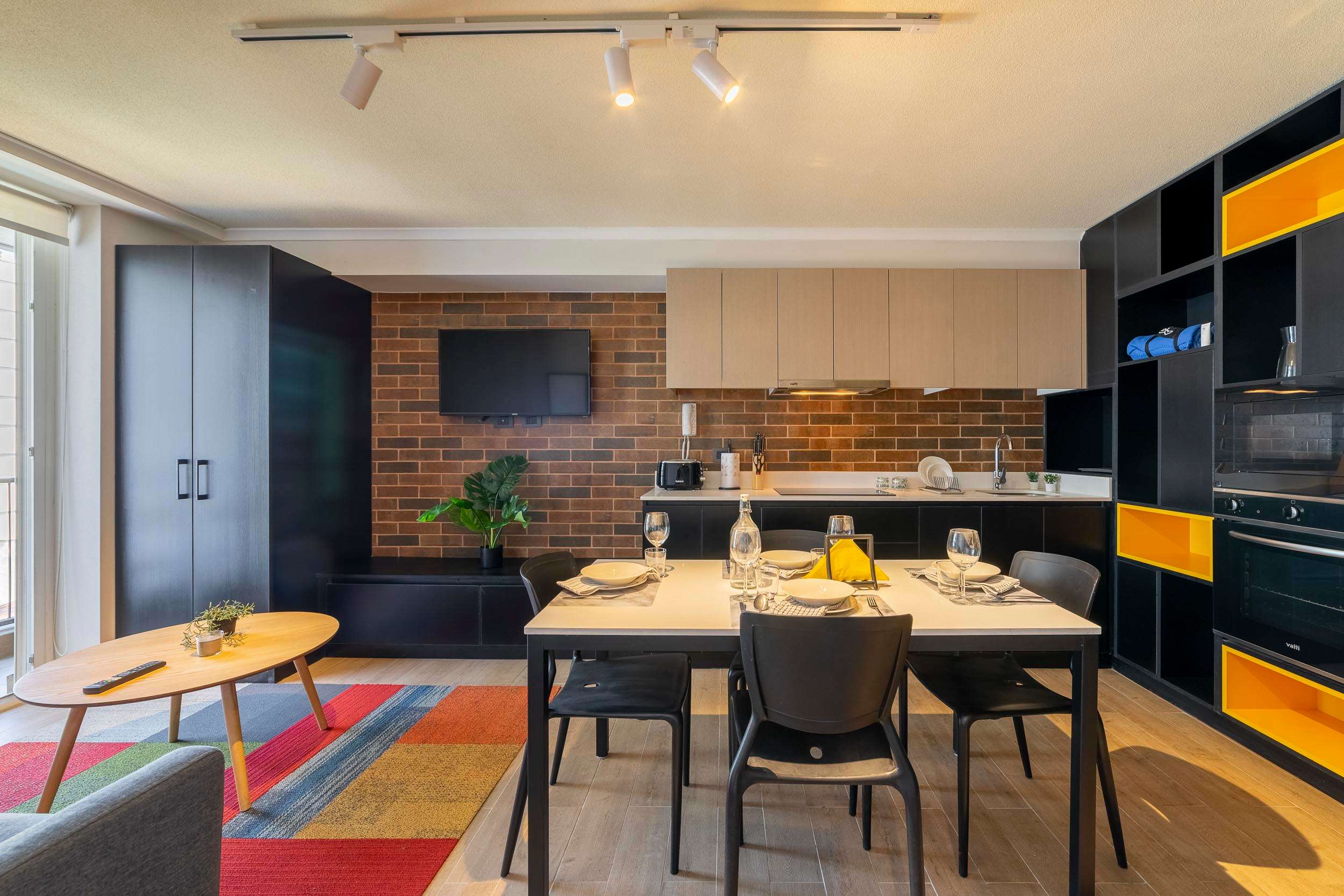 Private kitchen and dining area with an exposed-brick backsplash, modern black cabinetry, and a colorful geometric rug.