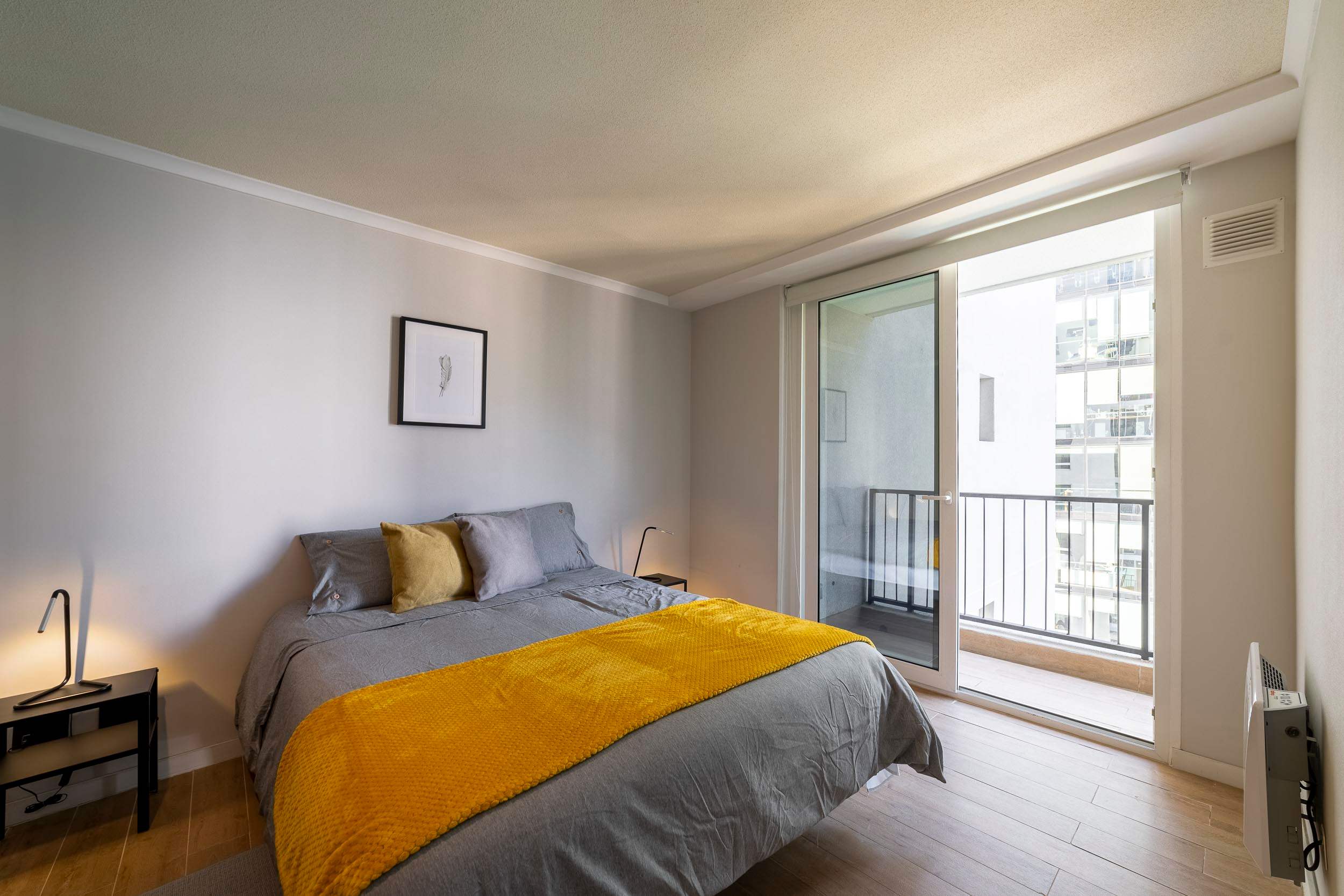 Bright studio bedroom with a cozy mustard throw and floor-to-ceiling doors opening onto a private balcony.
