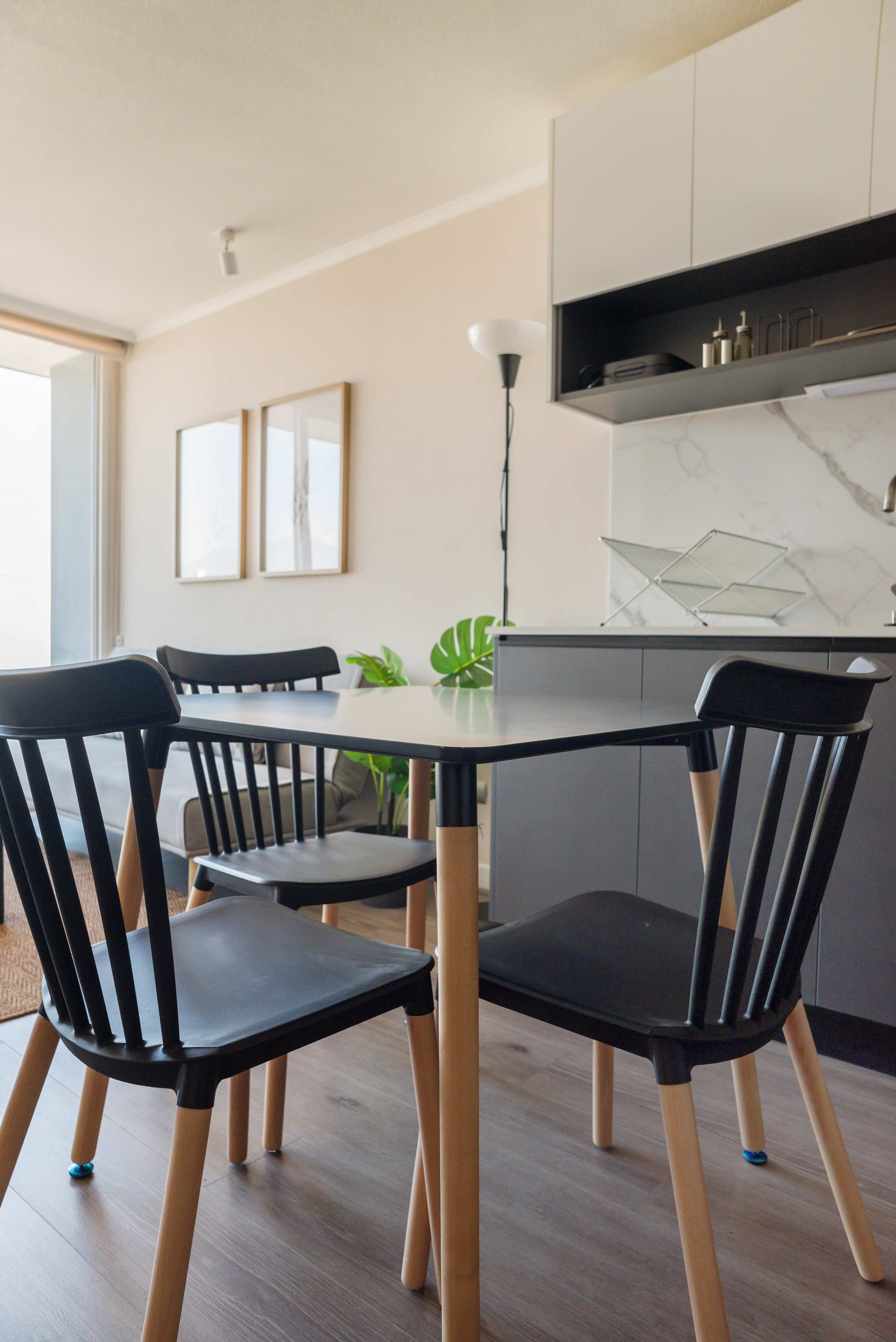 Bright dining area with a light wood table, black chairs, and a modern kitchen island accented by leafy green plants.