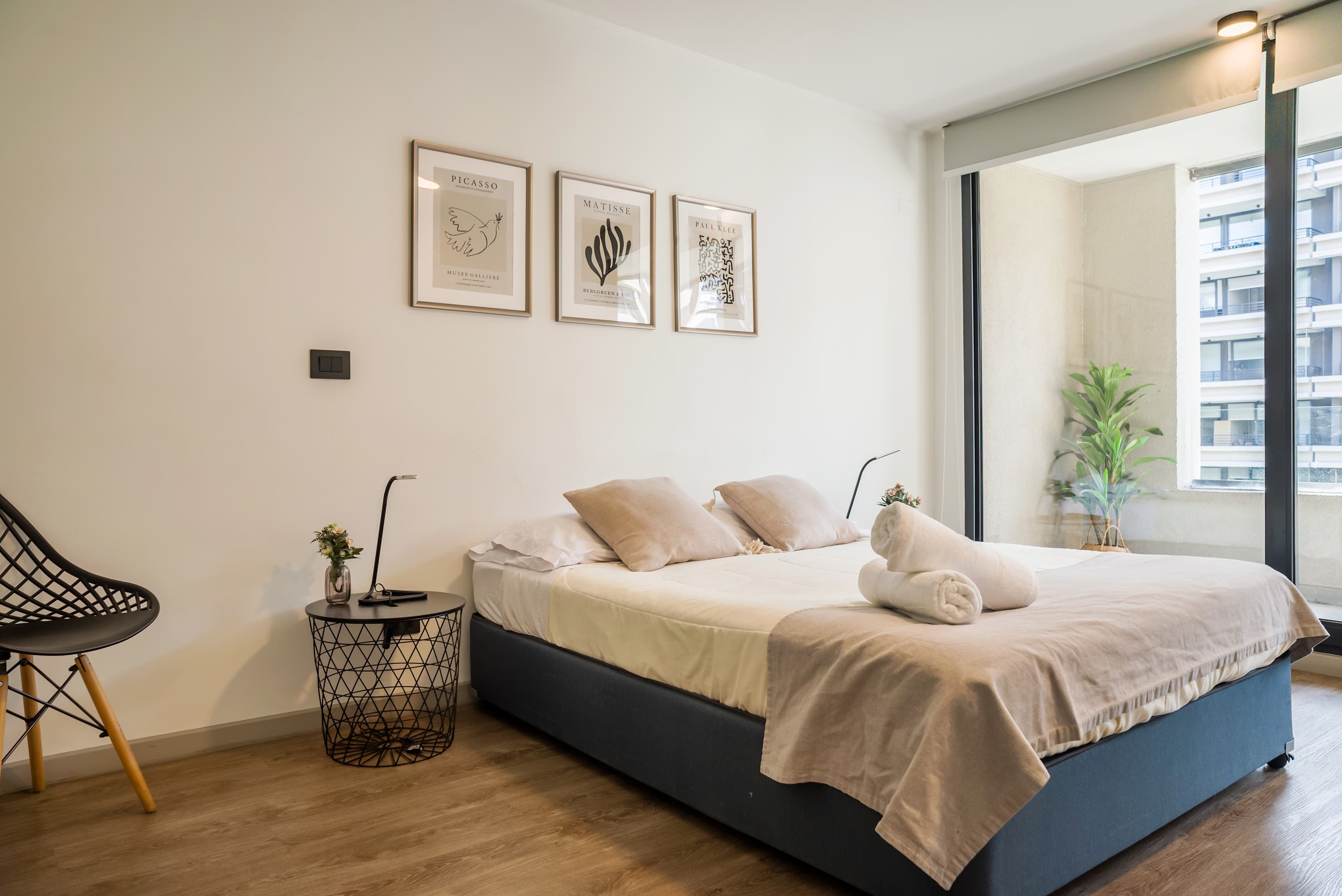 Bright bedroom with crisp white linens, rolled towels, contemporary art, and a large window overlooking the surrounding city buildings.
