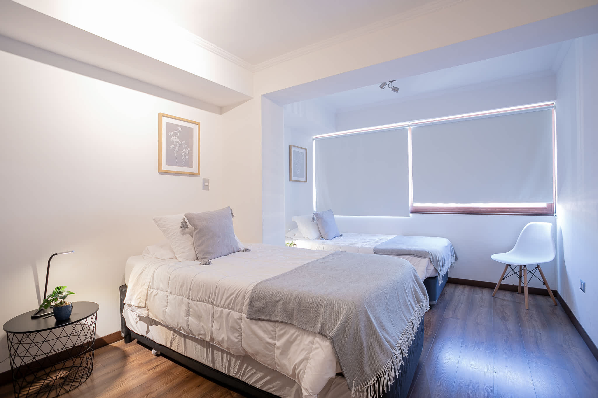 Bright private bedroom with two neatly made beds, soft gray throws, warm wood floors, and large windows with roller blinds.