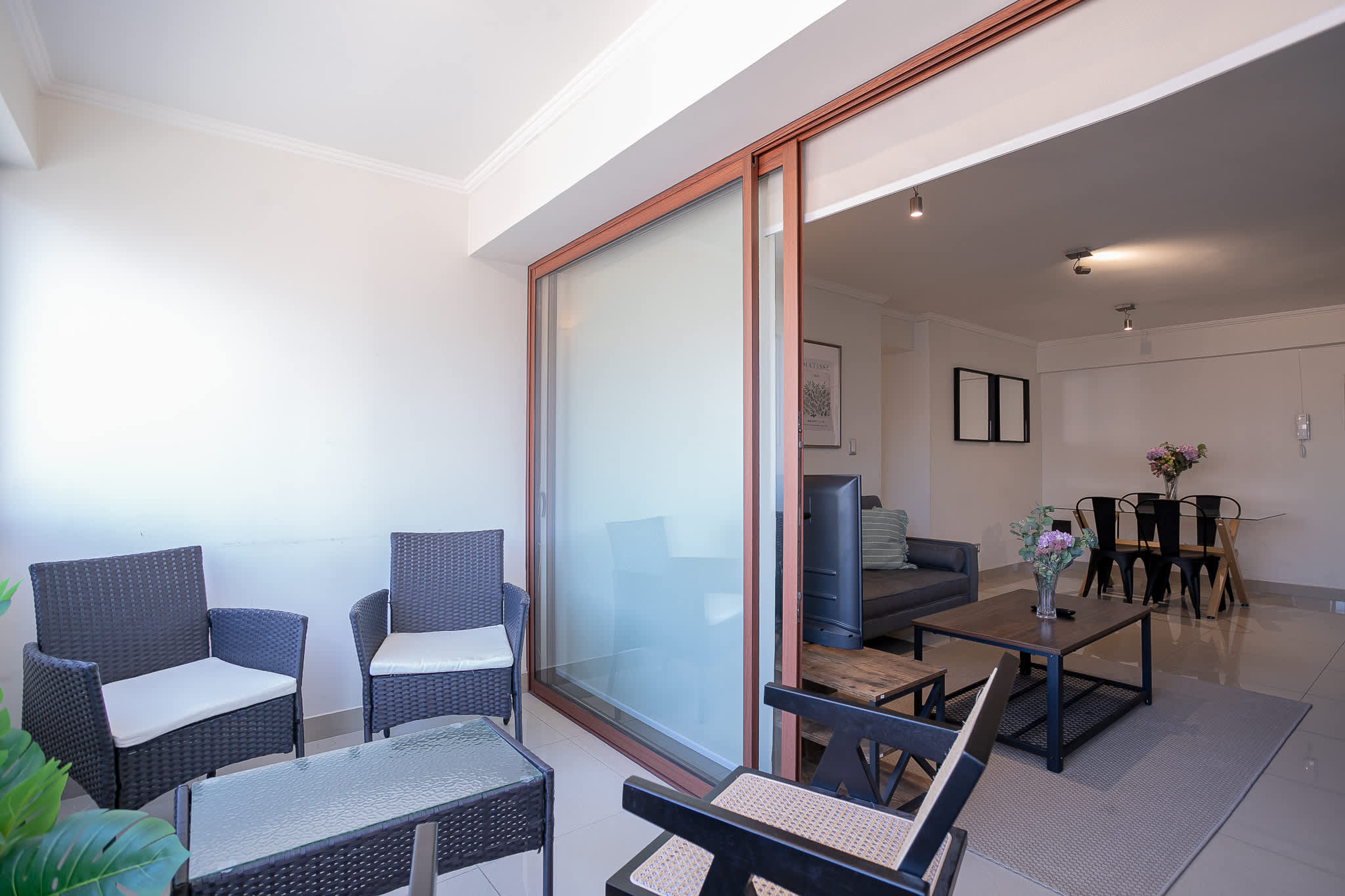 Bright living and dining area with a wood-framed glass partition, woven armchairs, dark sofa, and a flower-topped dining table.