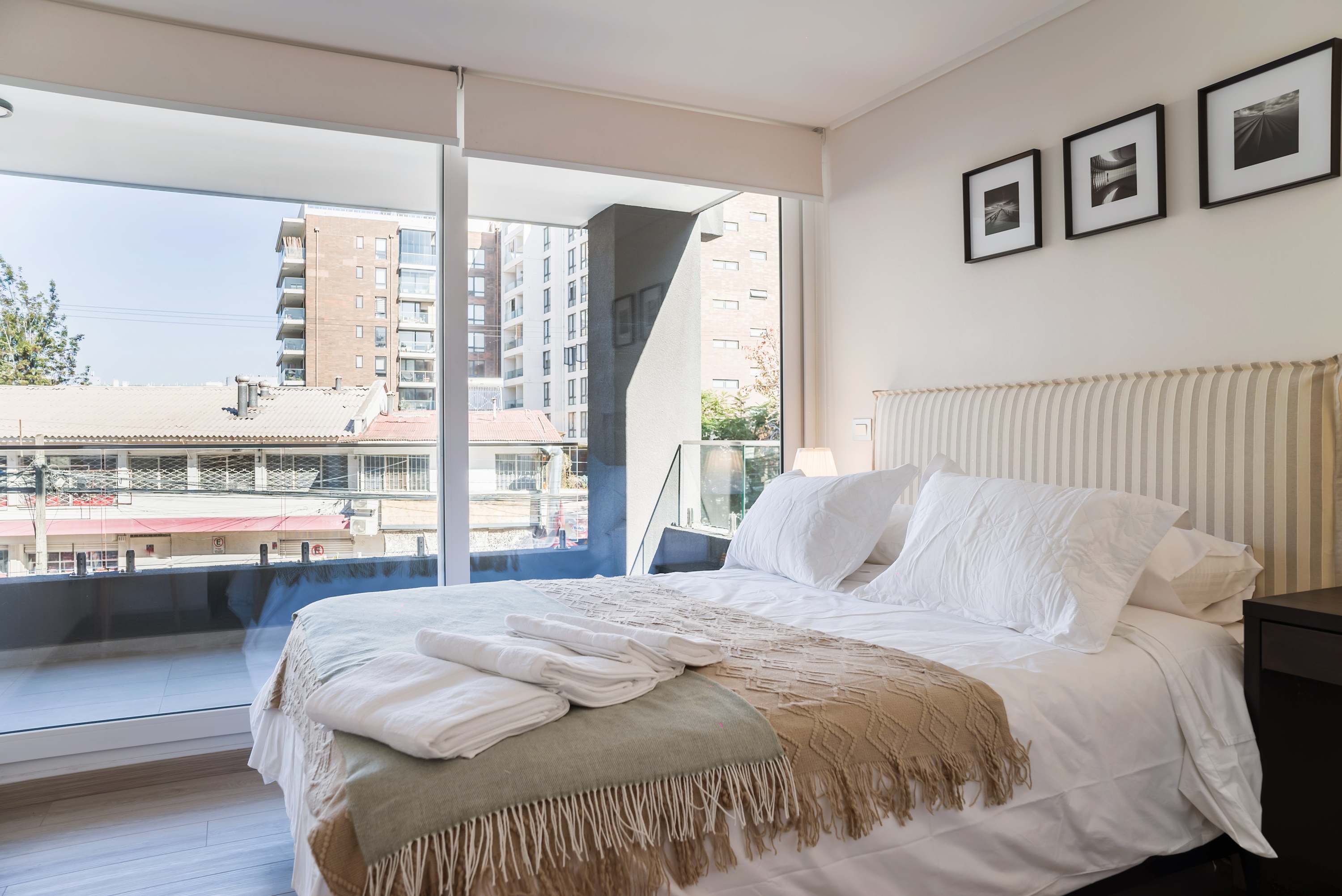 Bright bedroom with crisp white linens, a cozy textured throw, and floor-to-ceiling windows overlooking the surrounding city.