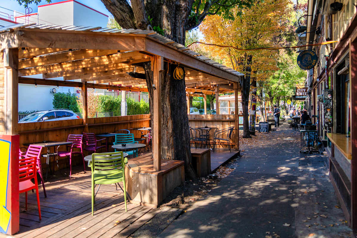 A wooden patio with colorful chairs and a pergola on a street lined with trees and buildings.