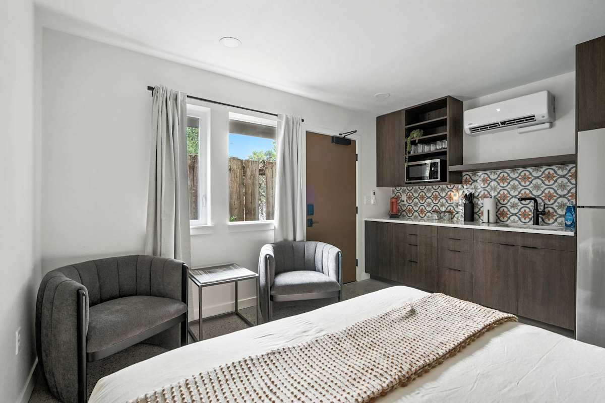 A wide view of a hotel room with a kitchenette, sofa, armchair, and bed.