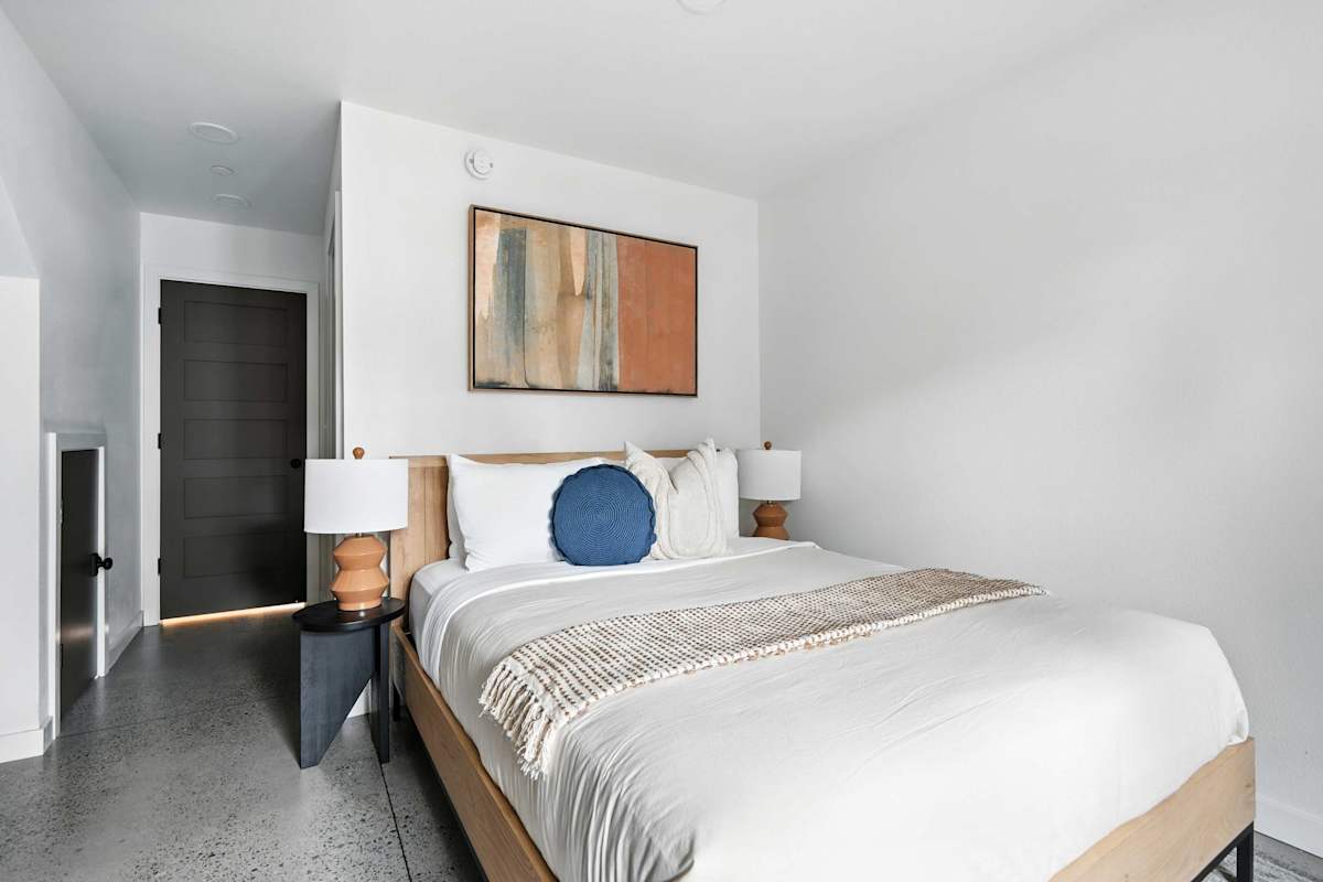 A neatly made bed with white linens and a blue accent pillow in a modern room with polished concrete floors and abstract artwork.