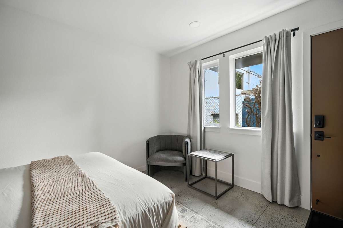 A white-walled guest room with a bed, chair, table, and window.