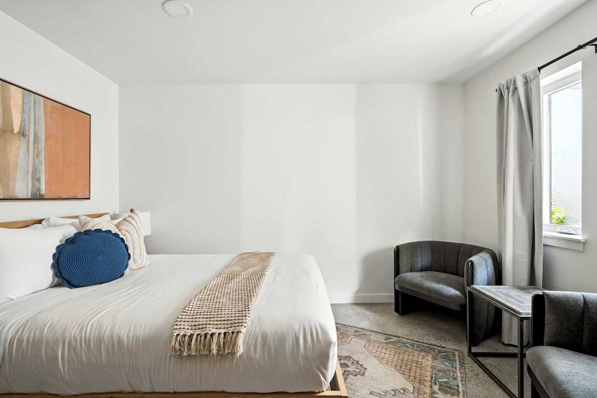 A neatly made bed with white linens and a blue accent pillow in a room with a window, two chairs, and a small table.
