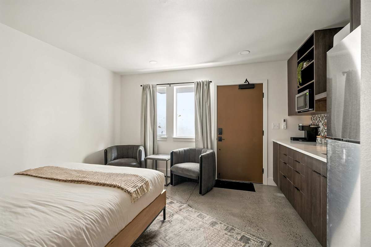 A hotel room with a bed, chair, window, and kitchenette.