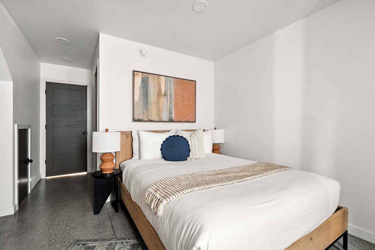 A neatly made bed with white linens and a blue accent pillow in a modern bedroom with a doorway to another room.