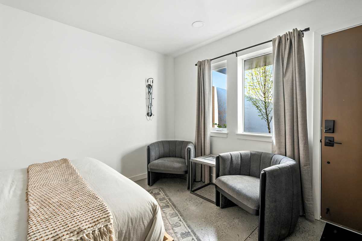 A hotel room with a bed, two chairs, a window, and a door.