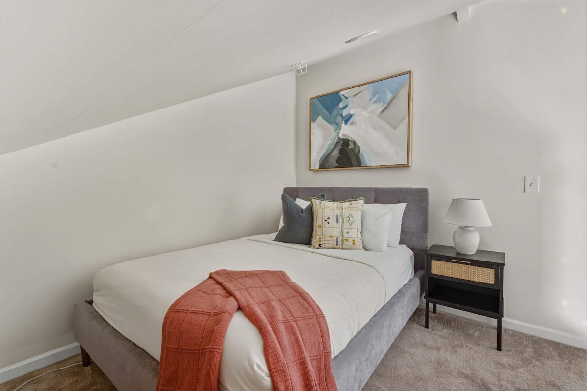 A neatly made bed with pillows and a blanket in a white room with a painting on the wall.