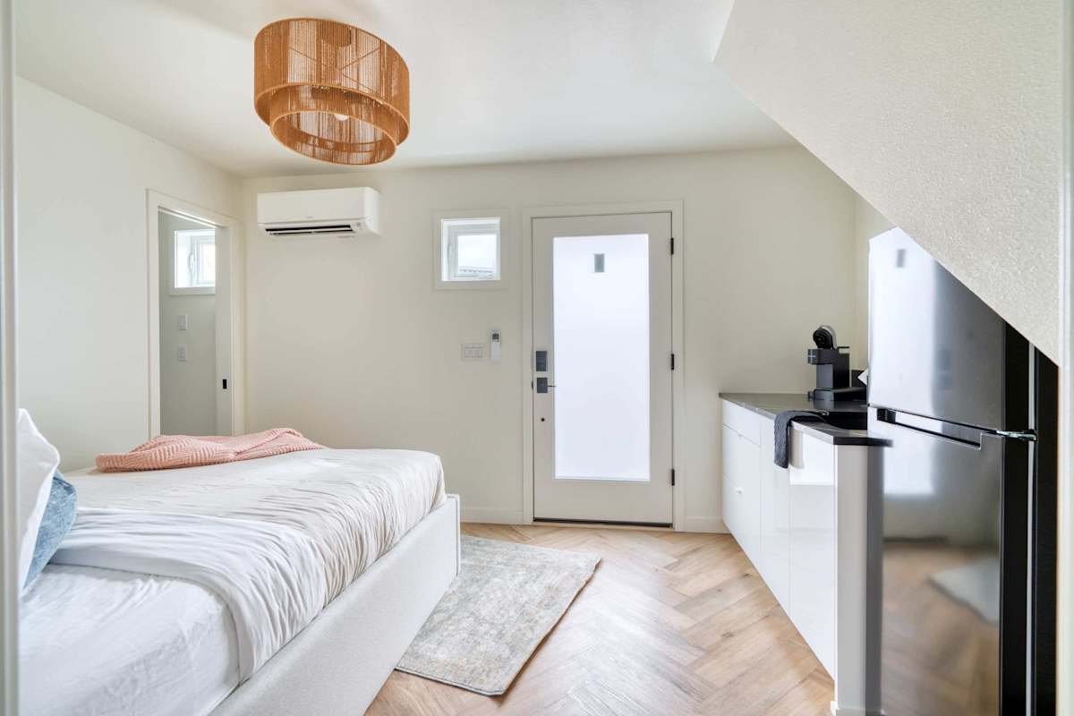 A small, white bedroom with a bed, TV, kitchenette, door, and window.