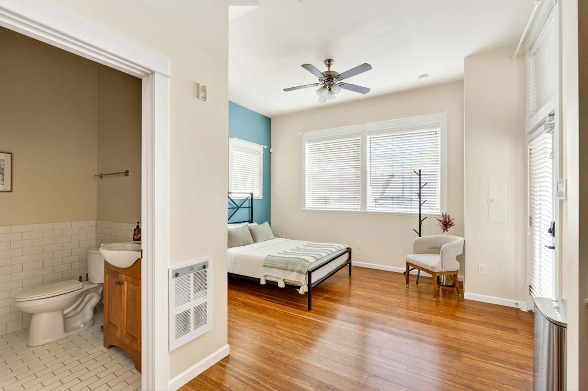 A wide view of a room with a bed, chair, and ceiling fan, with a bathroom in the background.