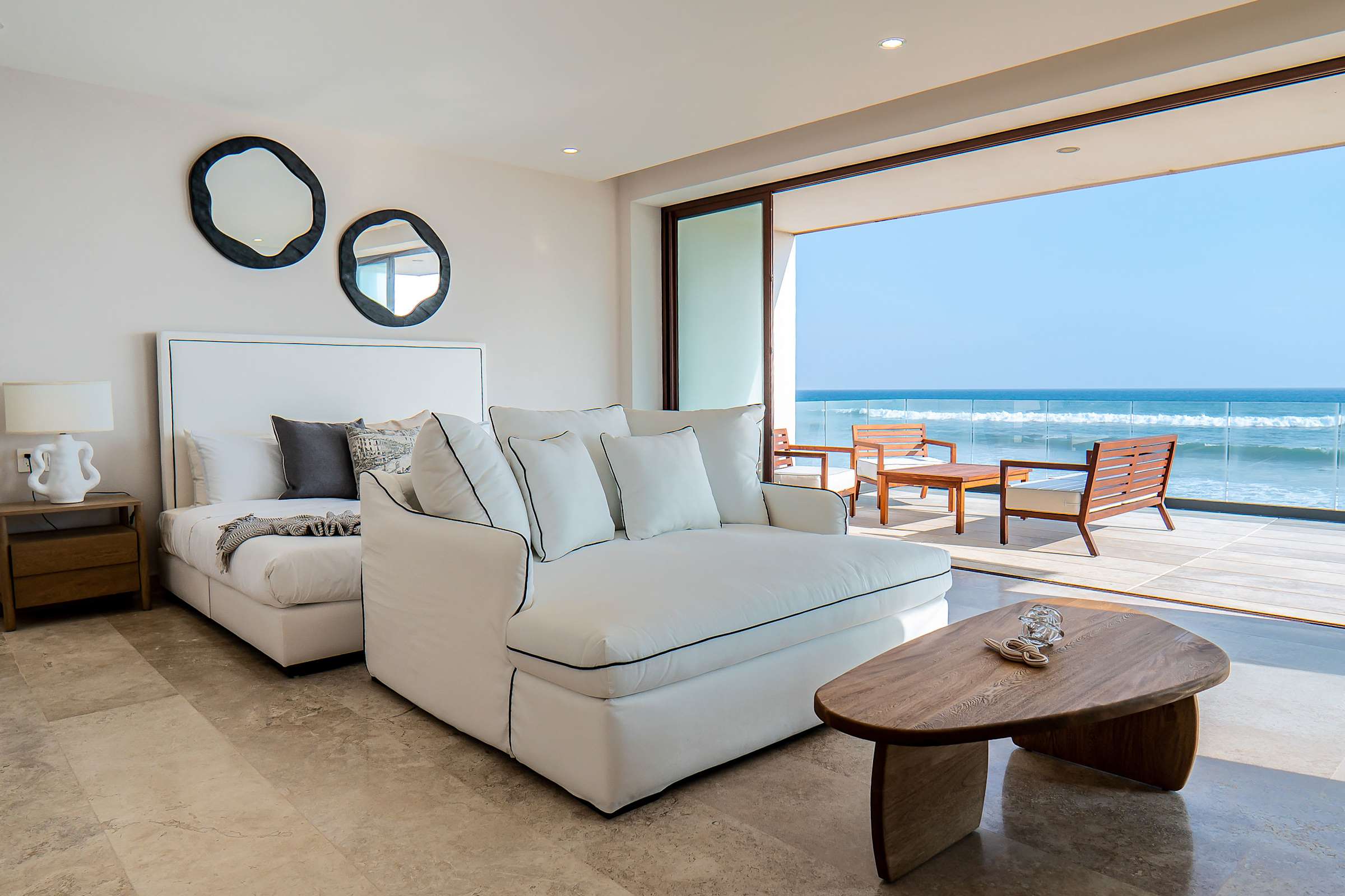 King suite sitting area with plush sofas, circular mirror wall art, and sweeping ocean views through glass walls