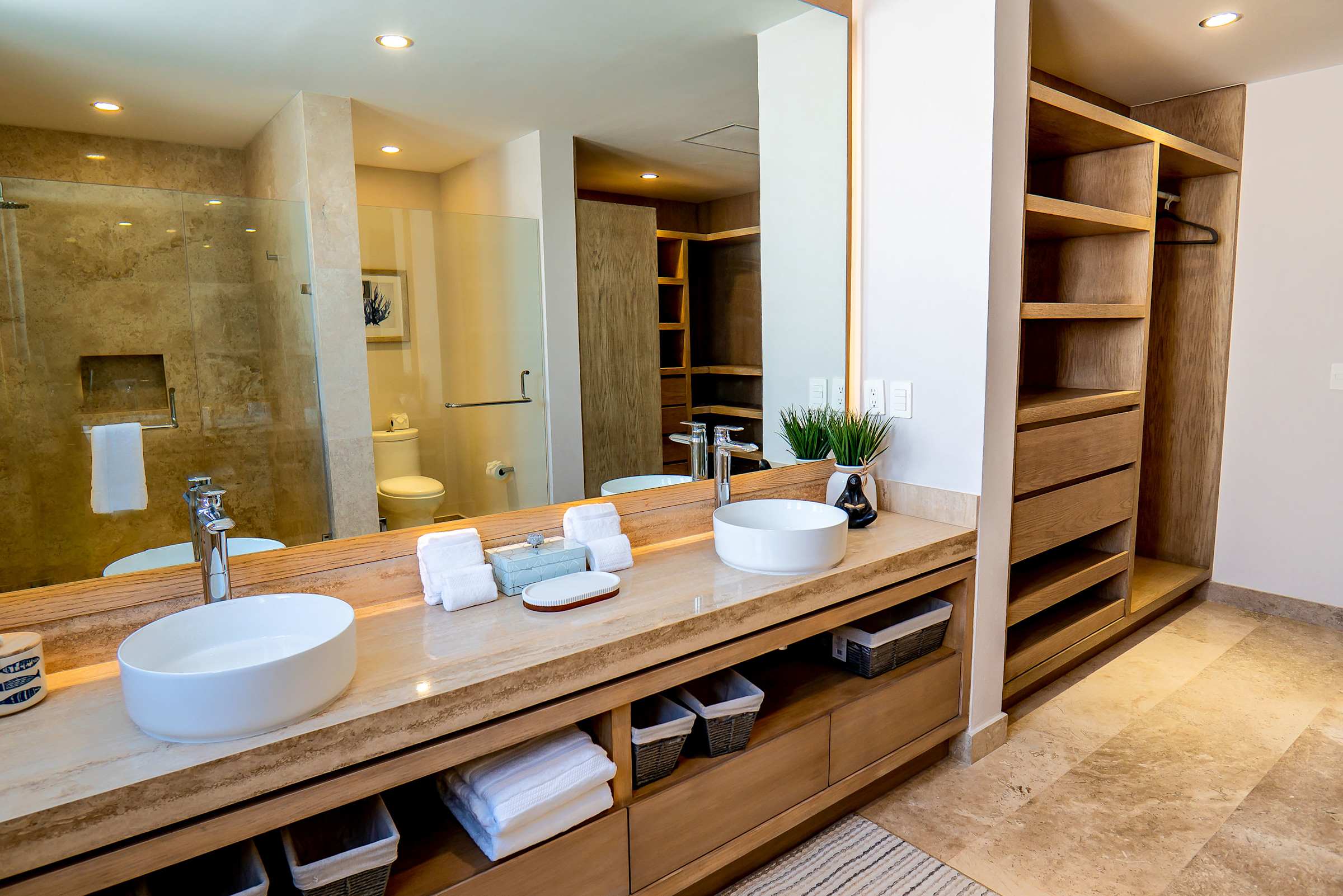 En-suite bathroom with double vanity sinks, warm wood cabinetry, and natural light — a spa-like retreat. Includes wood shelving and premium bath linens.