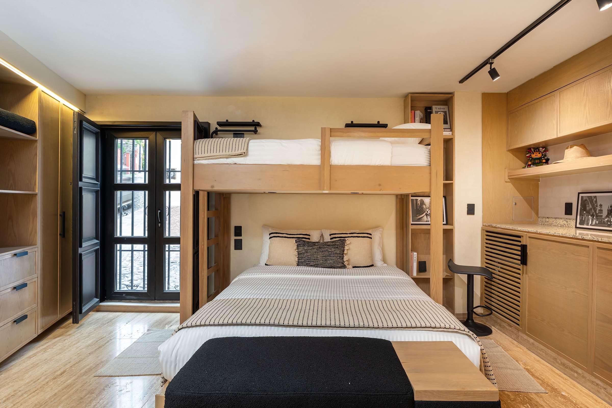 Bedroom featuring a queen bed with a twin bed above, large black-framed windows, open wardrobe storage, and wood built-in shelving along the wall.