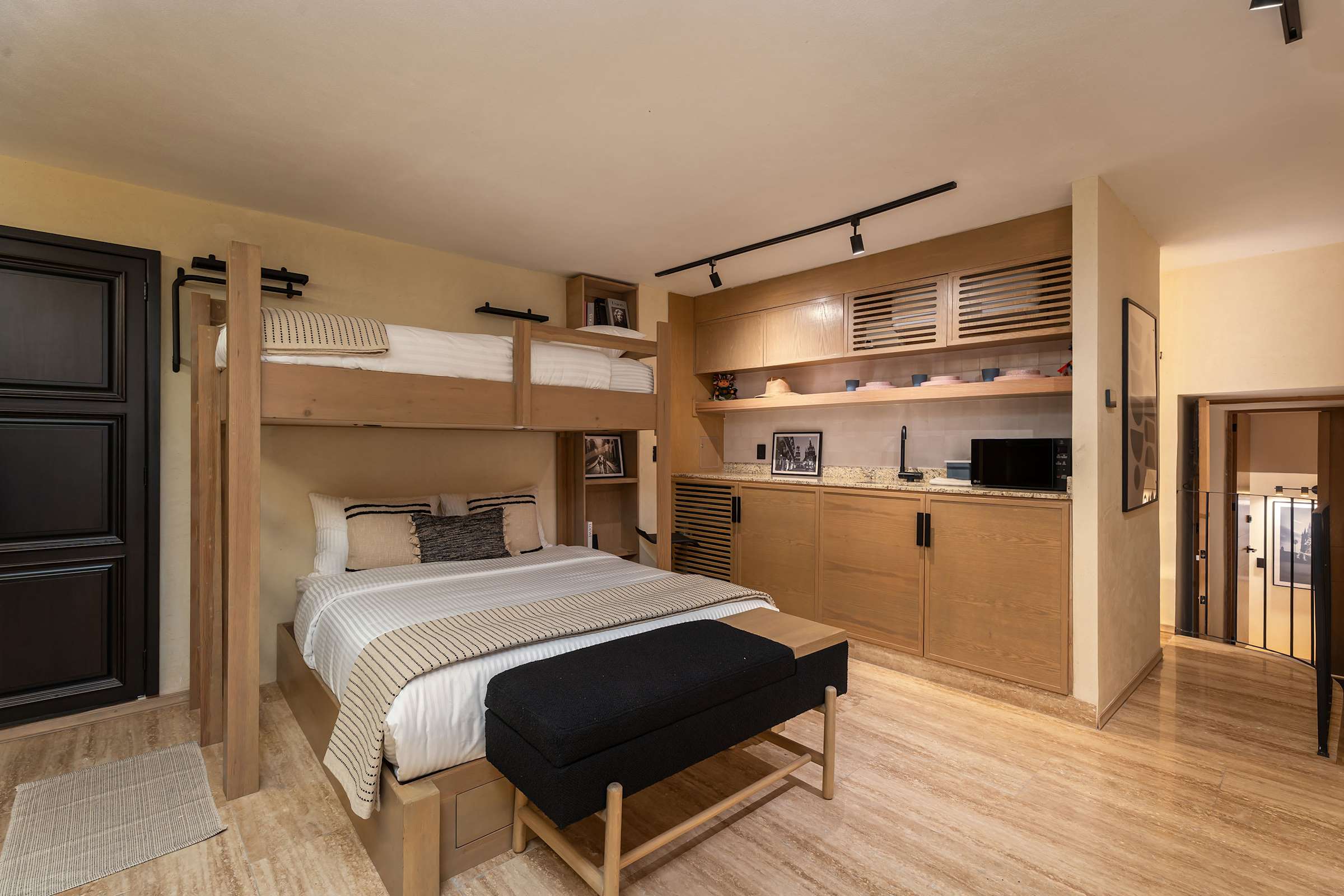 Bedroom with a queen bed below a twin loft bed, extensive wood built-in storage and shelving, a kitchenette with microwave, and a hallway leading to the bathroom and other room.
