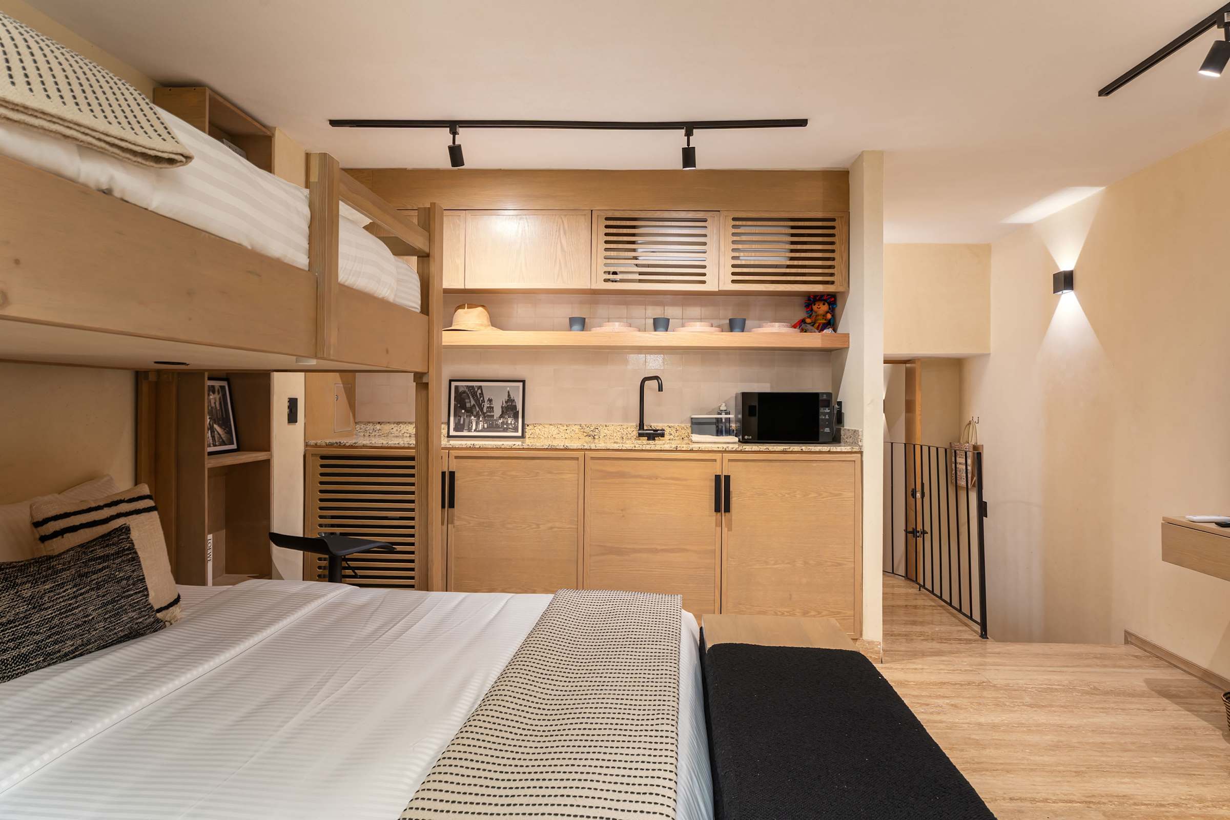 Bedroom detail showing the built-in twin loft above the queen bed, warm wood shelving with decorative items, and the kitchenette area with a microwave.