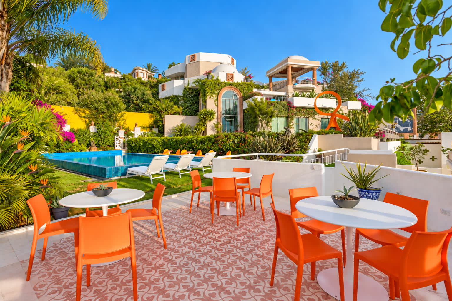 Terraza con sillas naranjas, mesas de comedor, alberca y jardines con plantas tropicales